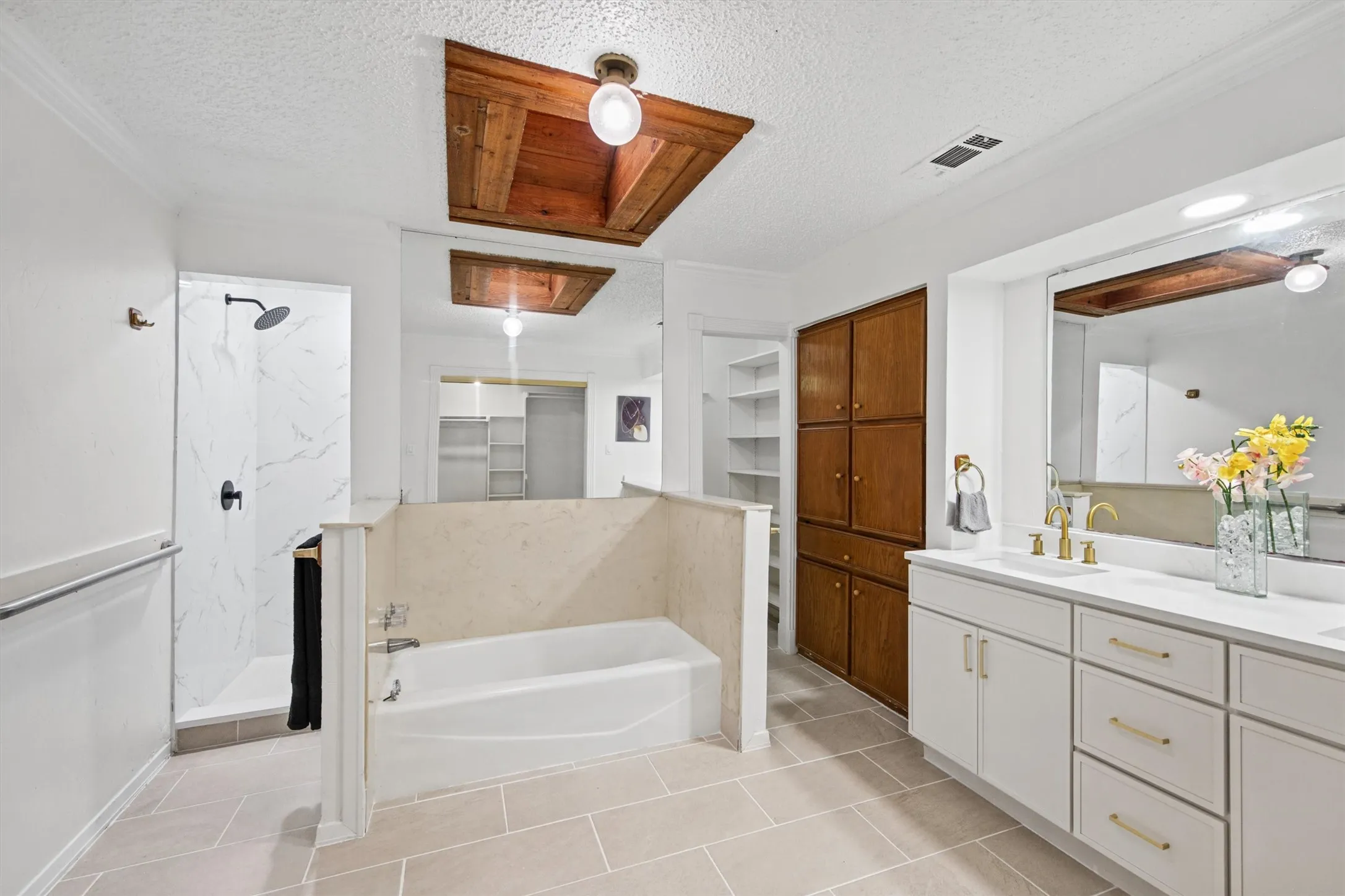 Tiled flooring, white built-in double vanity with gold-tone fixtures, wood-tone built-in cabinetry, white soaking tub, and a separate shower with marble-look wall panels