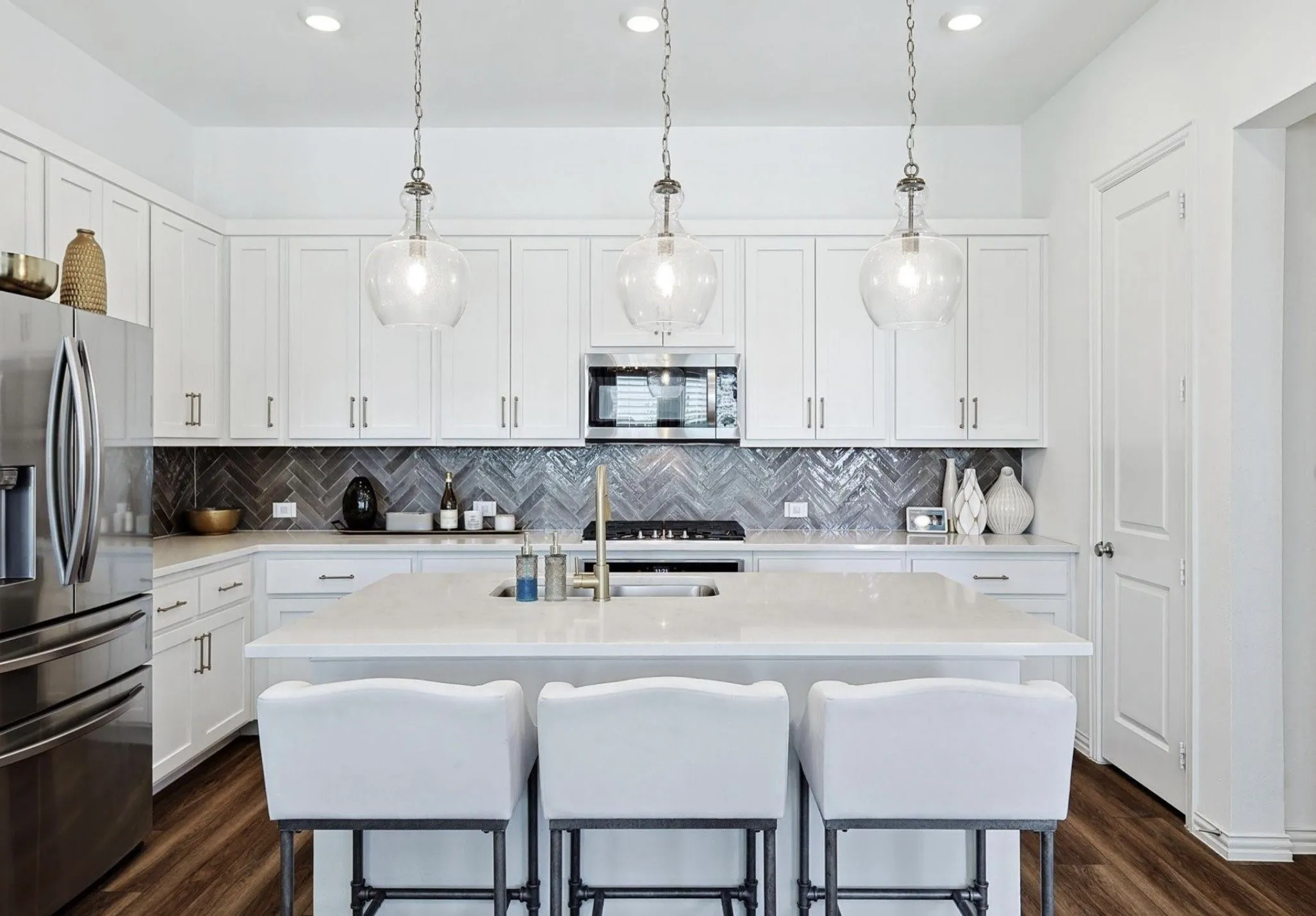 White cabinetry with brushed nickel hardware, stainless steel appliances, herringbone tile backsplash, an island with a light-colored countertop, and wood-finish flooring