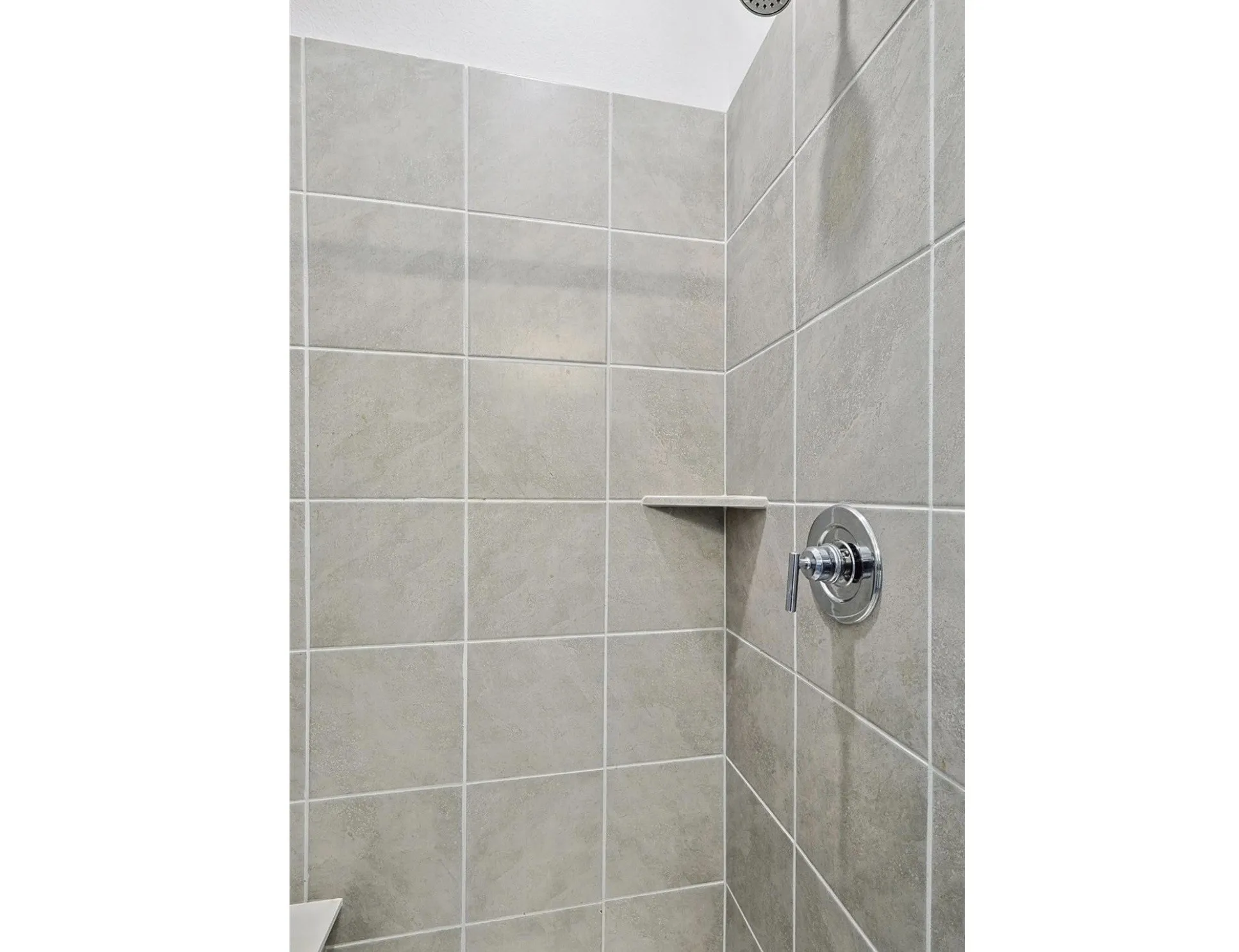 Light grey square wall tiles with white grout, polished chrome shower control, and a built-in shower shelf