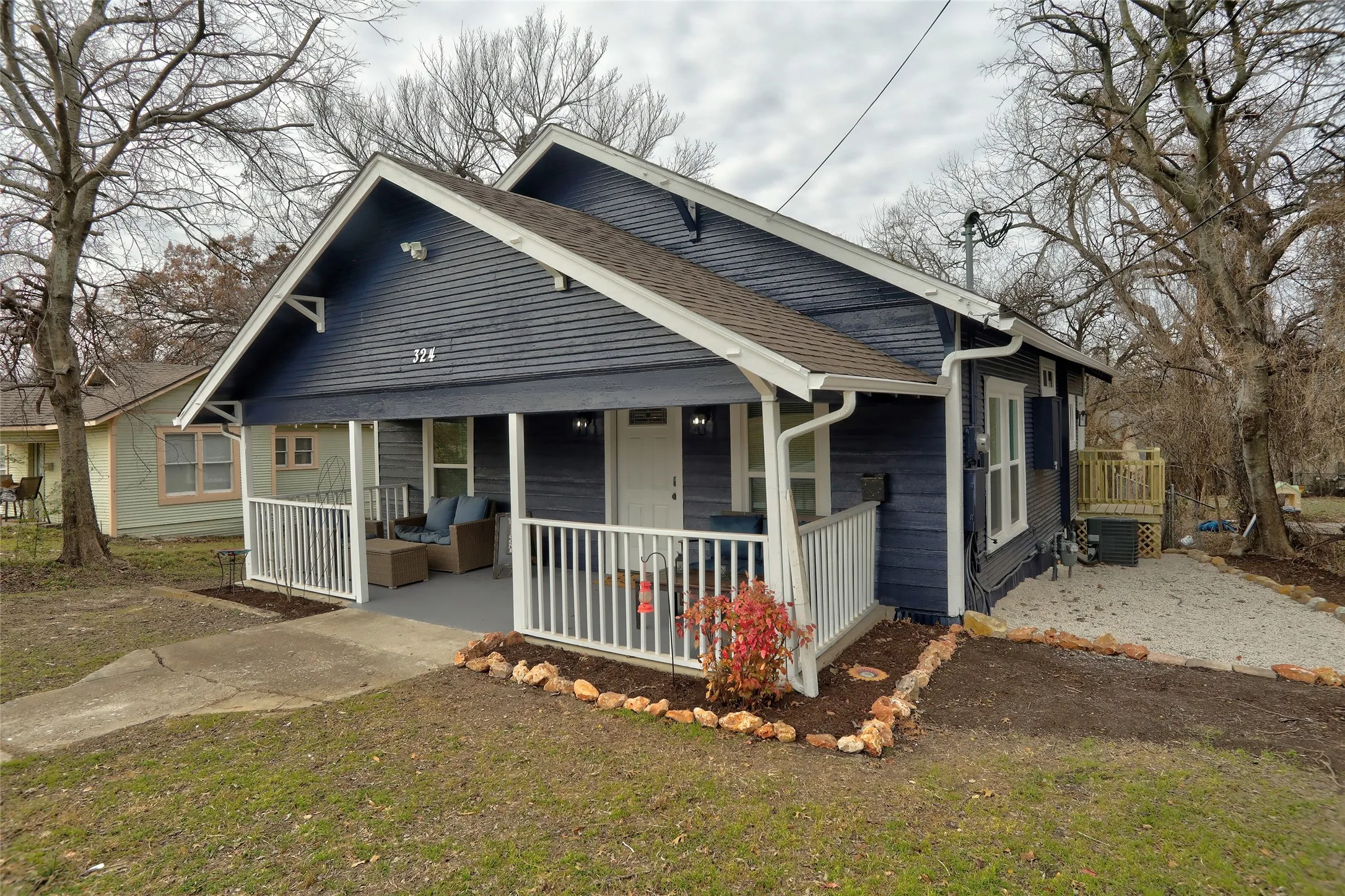 Exterior featuring dark blue siding, a covered front porch with white railing, a side deck, and a landscaped front yard with decorative rock and mulch
