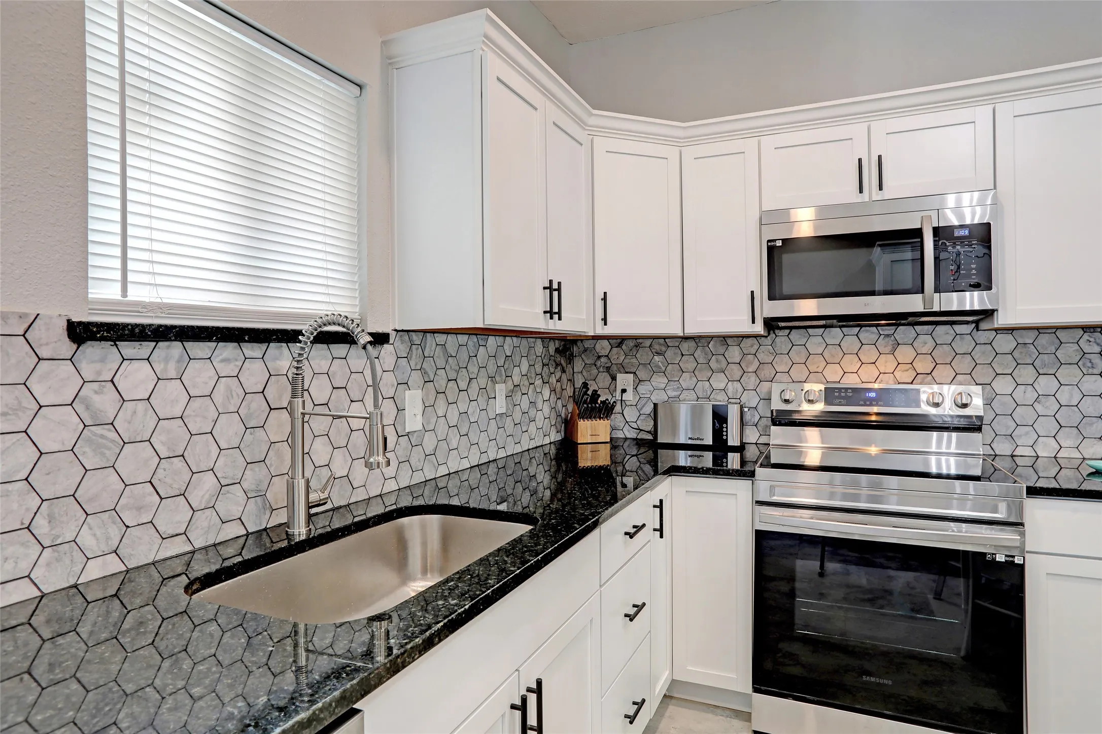 White shaker cabinetry with crown molding, dark countertops, hexagonal tile backsplash, stainless steel appliances, and a stainless steel sink with a pull-down faucet