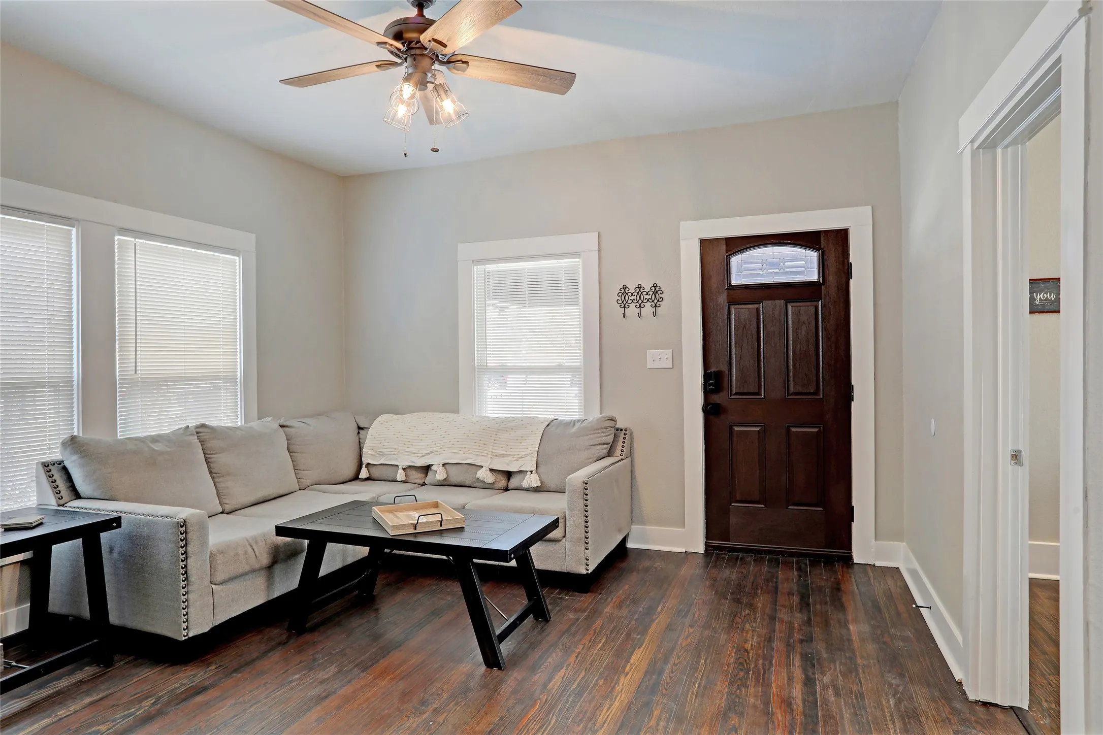 Rich wood-finish flooring, a dark wood paneled entry door with transom, a ceiling fan with integrated lighting, and multiple windows with blinds