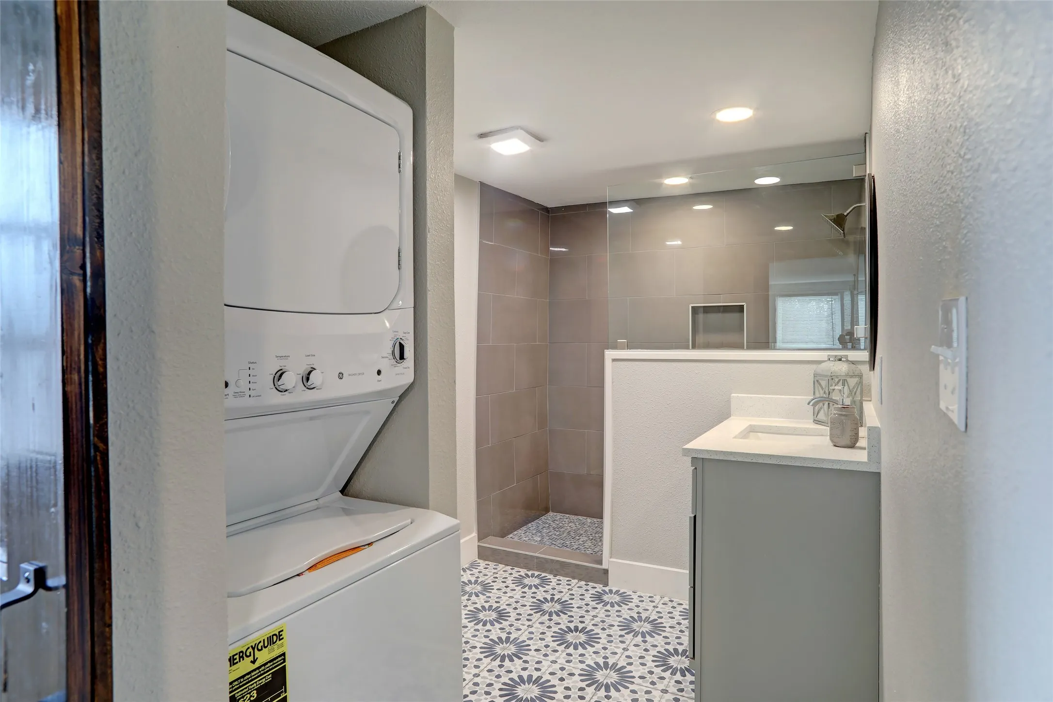 Stackable washer and dryer, vanity with a white countertop and integrated sink, glass-enclosed shower with a tiled surround, recessed lighting, and patterned floor tiling