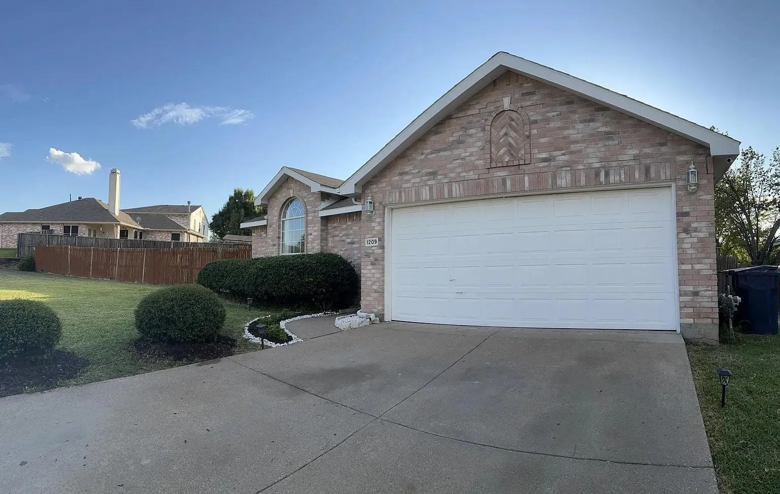 Brick exterior with an arched window, attached garage, concrete driveway, and established landscaping