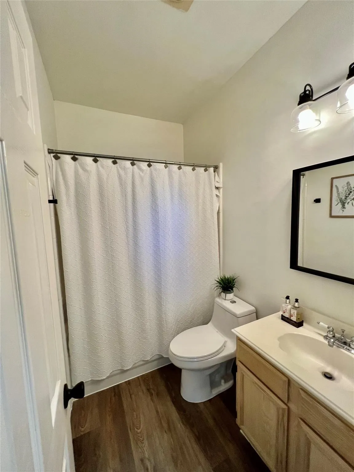 Bathroom featuring wood-finish flooring, a single vanity with light-tone cabinetry, a neutral countertop, and a shower with a white curtain