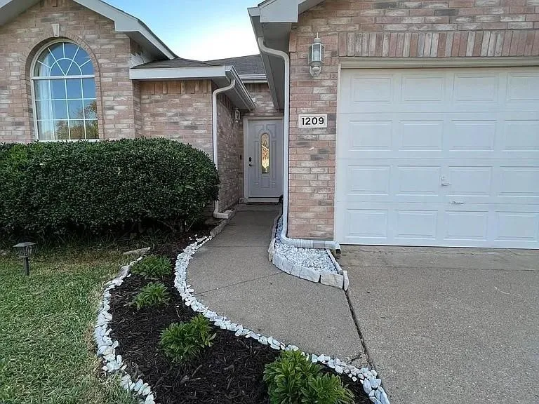Brick facade with an arched window, white paneled garage door, concrete driveway, and a landscaped walkway with white stone accents