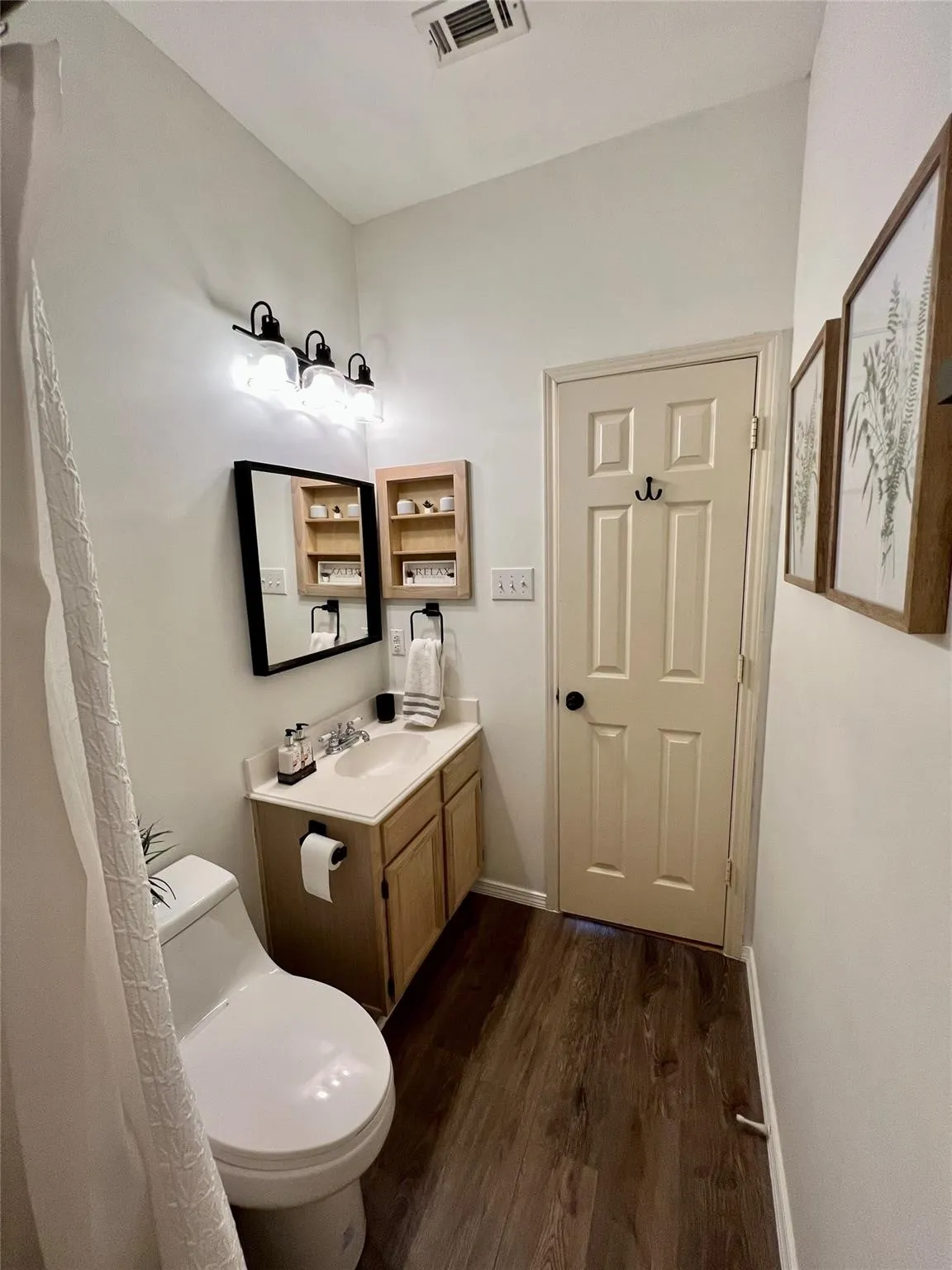 Wood-finish flooring, single vanity with light-tone cabinetry, wall-mounted light fixture, and a framed mirror