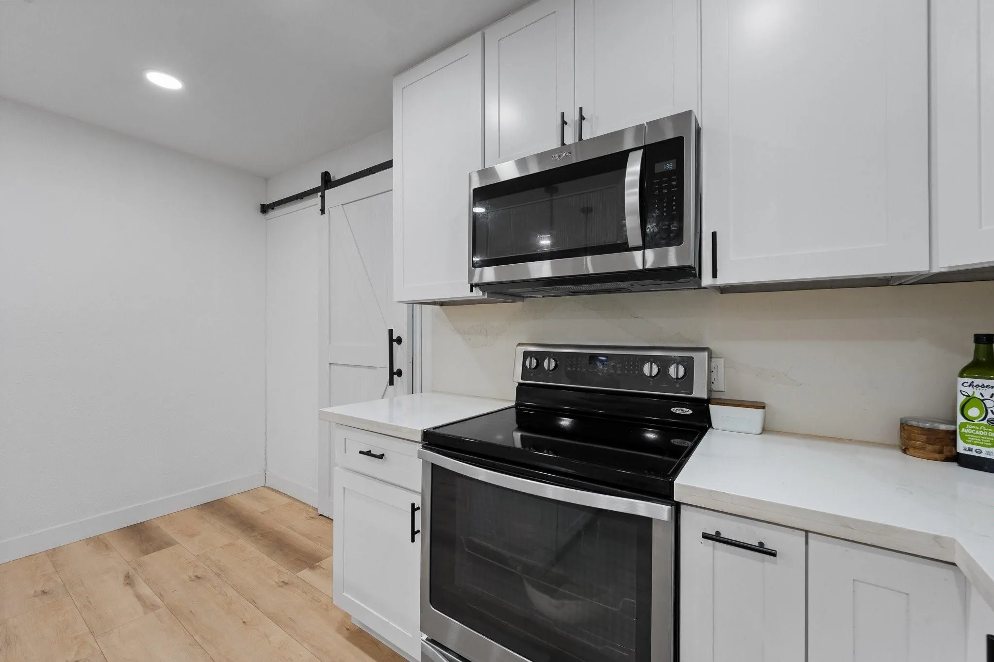 Stainless steel appliance suite, white shaker cabinetry, light-toned wood-finish flooring, white countertops, and a barn door