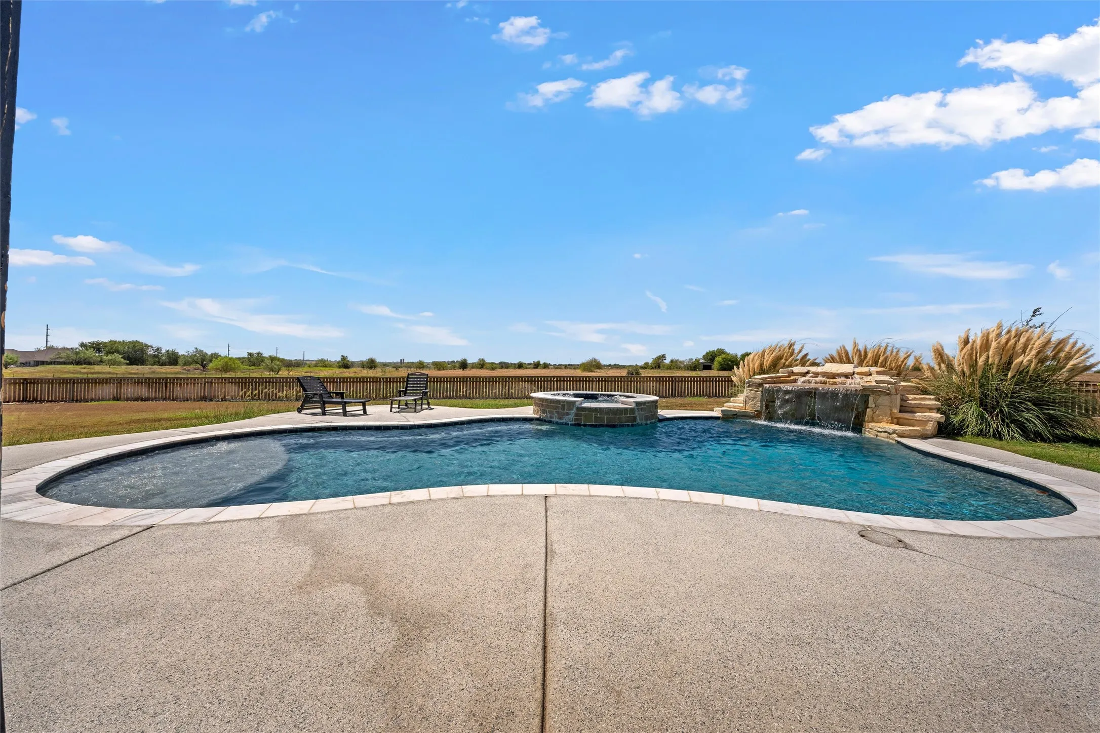 Curved swimming pool with integrated spa, concrete deck, rock waterfall feature, and surrounding landscaping