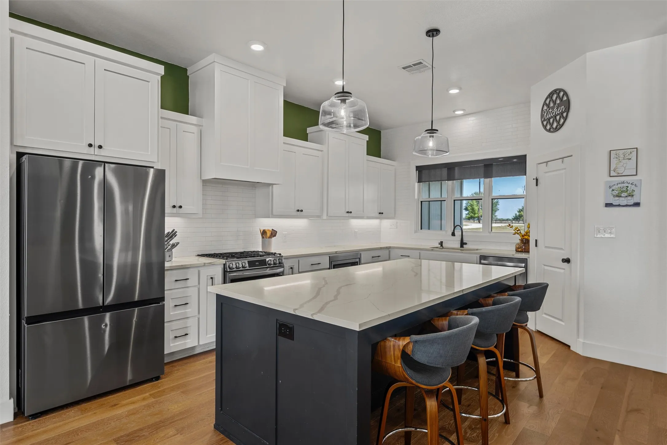 Kitchen featuring stainless steel appliances, white shaker cabinetry, an expansive island with a light-toned countertop, wood-finish flooring, and pendant lighting