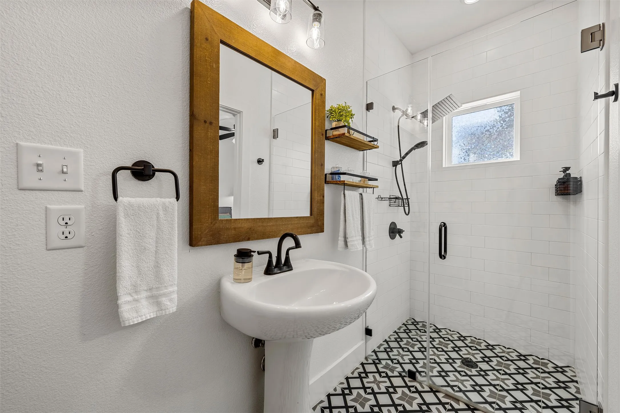 Pedestal sink with matte black faucet, framed mirror, glass-enclosed shower, shower with a rainfall head and handheld sprayer, and patterned floor tile
