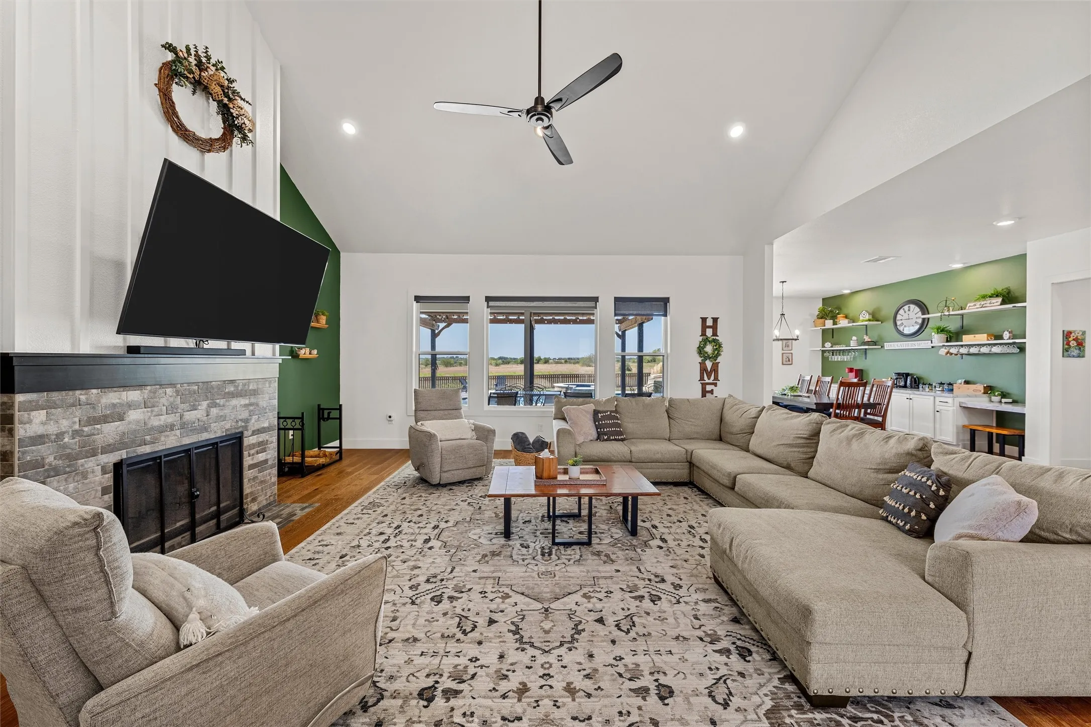 Vaulted ceiling with a ceiling fan, stone-clad fireplace with a dark mantel, wood-finish flooring, recessed lighting, and large windows