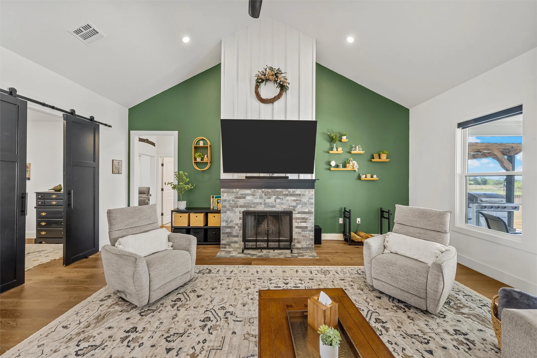 Vaulted ceiling, stone-clad fireplace, wood-finish flooring, accent wall, and sliding barn doors