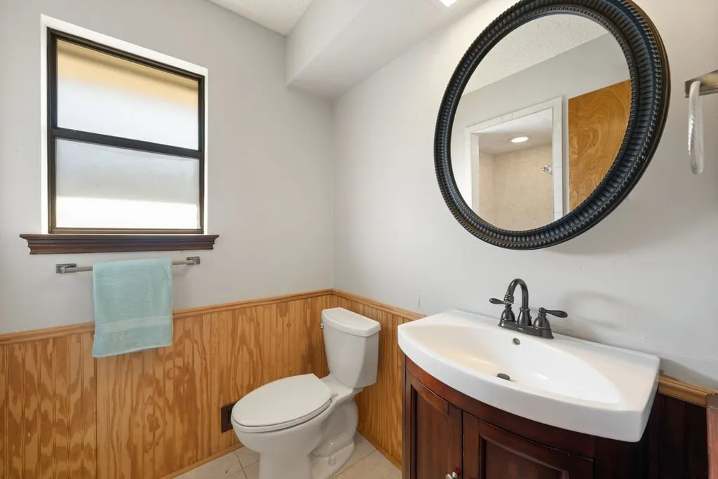 Powder room featuring a window, wood-finish wainscoting, a dark wood-finish vanity with an integrated sink, and an oval framed mirror