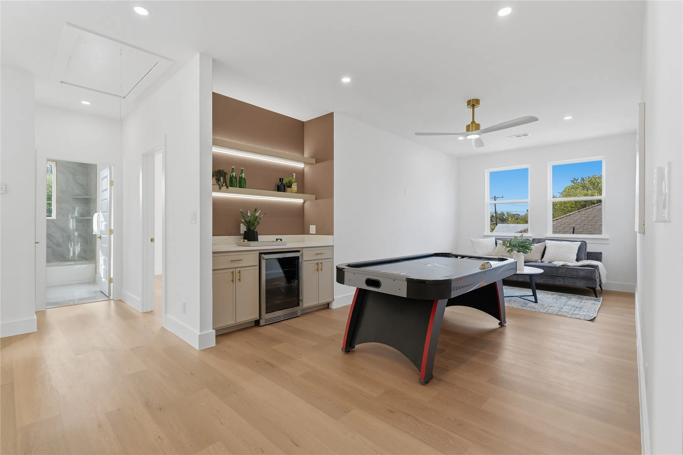 Light wood-finish flooring throughout, integrated wet bar with built-in beverage refrigerator, two large windows, recessed lighting, and a ceiling fan