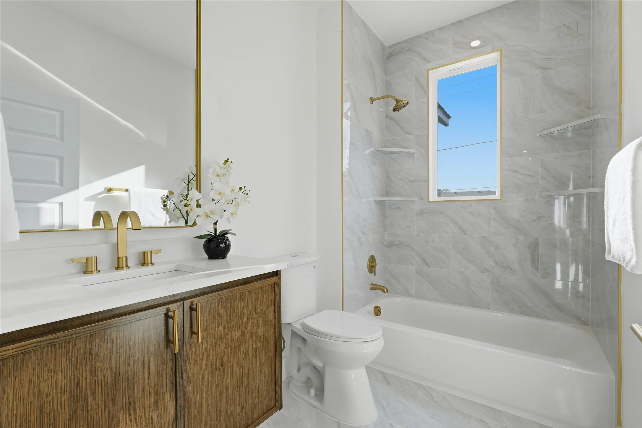 Bathroom with a wood-finish vanity, white countertop, gold-tone fixtures, and a white bathtub with tile surround
