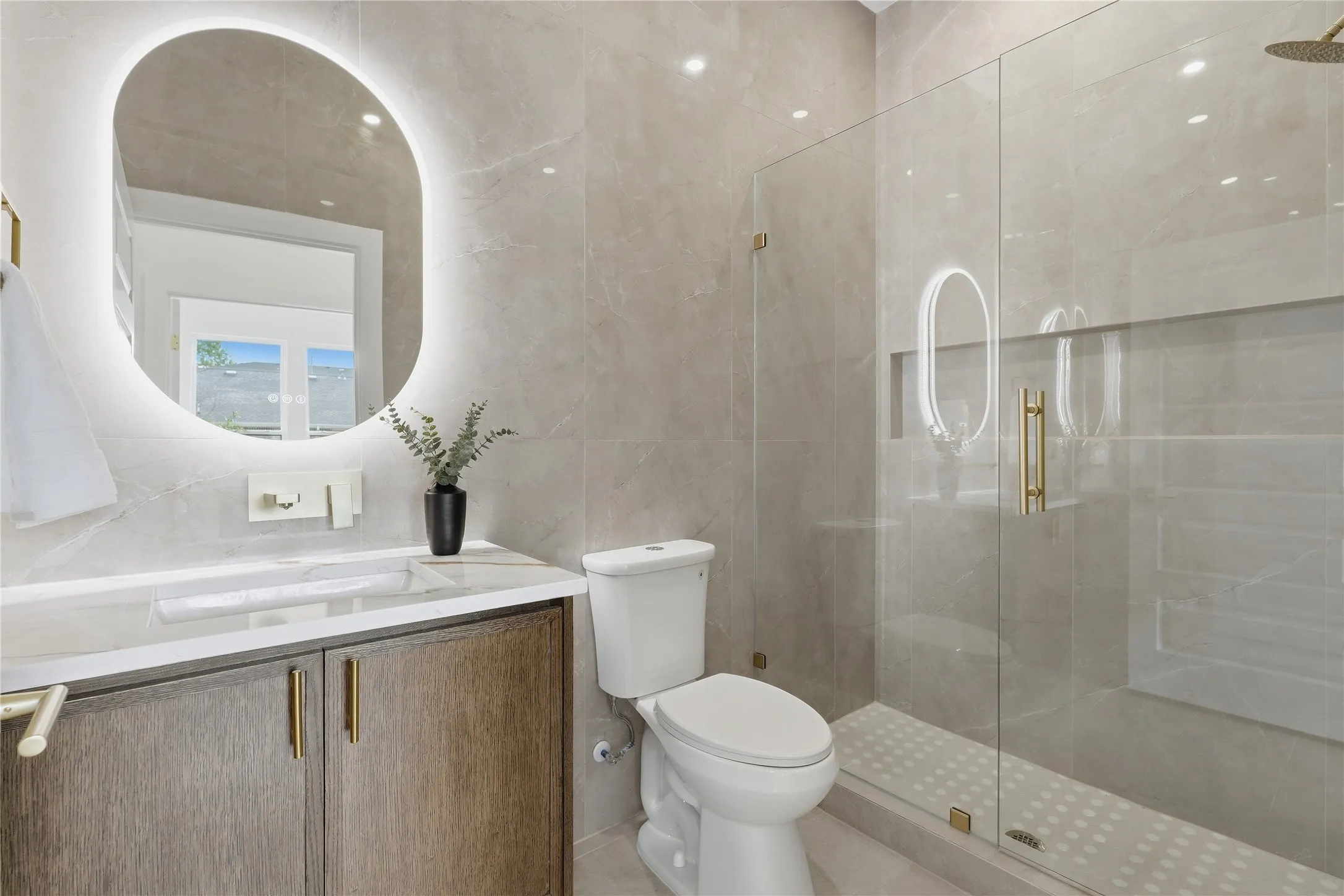 Illuminated oval mirror, wood-finish vanity with gold-tone hardware, white countertop with integrated sink, glass-enclosed shower, and full-height wall tiling