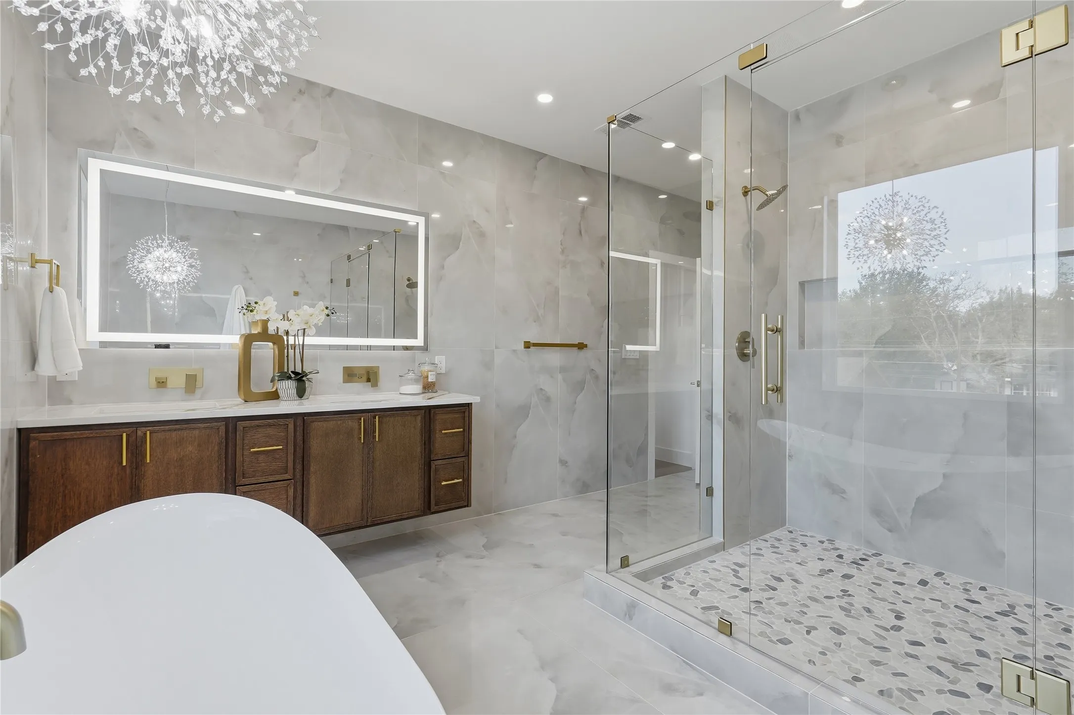 Freestanding soaking tub, dual vanity with wood cabinetry, backlit mirror, glass-enclosed shower with pebble-style floor, and ceiling-mounted light fixture