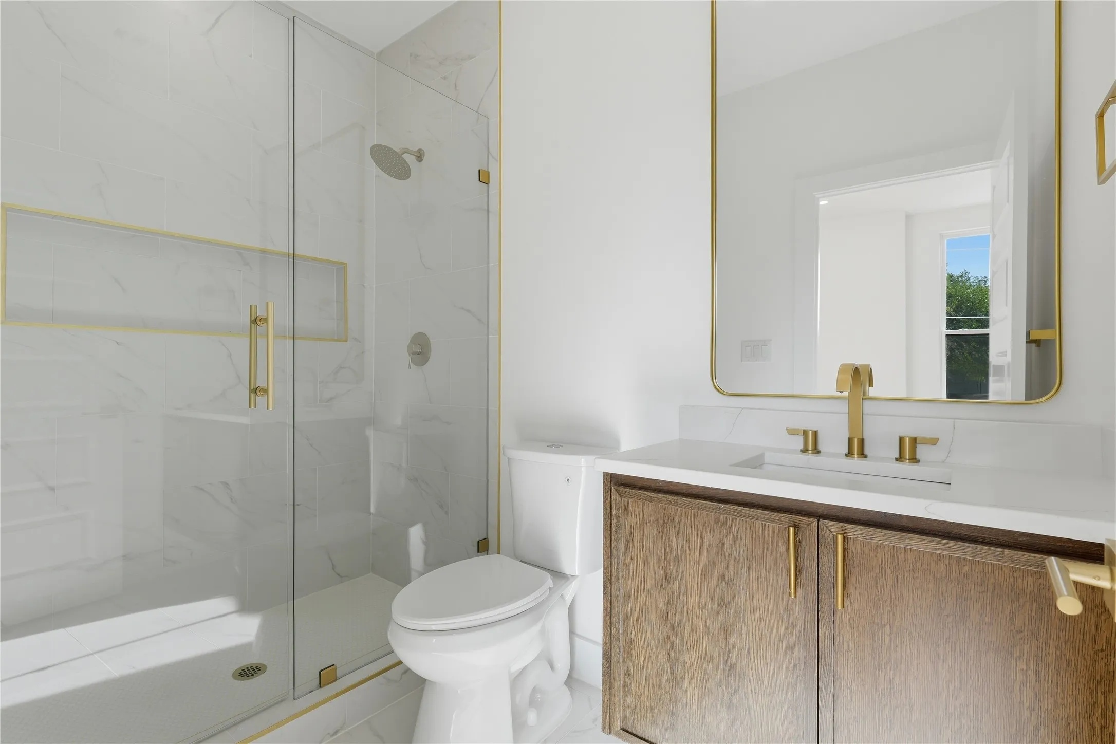 Walk-in shower with glass enclosure and gold-finish hardware, white tile with subtle veining, wood-finish vanity with gold-finish pulls, gold-finish faucet, and a rectangular mirror with a gold-finish frame