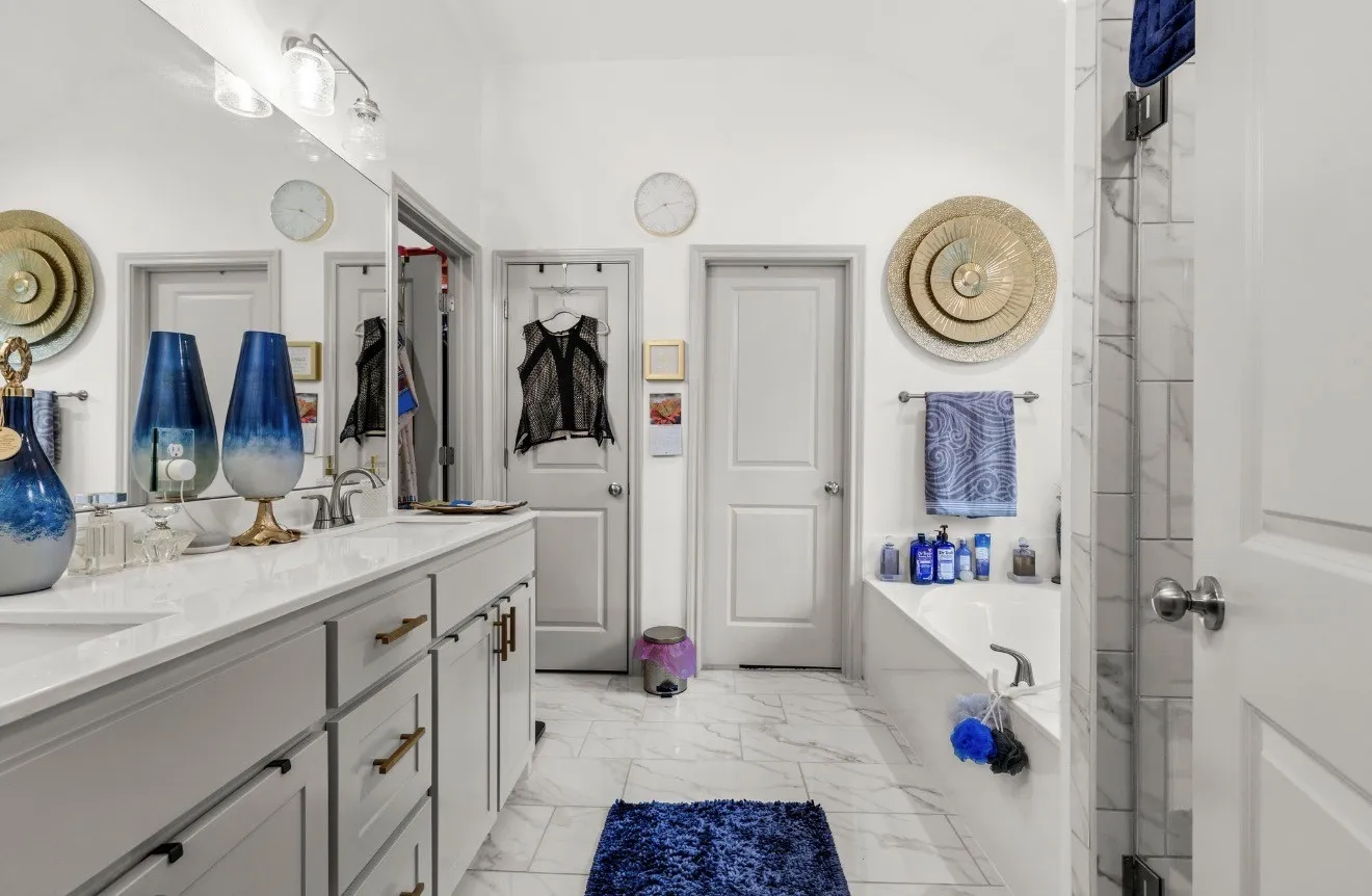 Dual vanity with white cabinetry and bronze hardware, light-toned countertop, large wall-mounted mirror, white tile flooring, and built-in bathtub with tiled surround