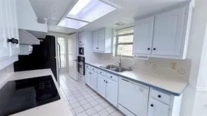 White cabinetry with black hardware, integrated kitchen appliances, dual basin sink, white tiled flooring, and a ceiling-mounted skylight