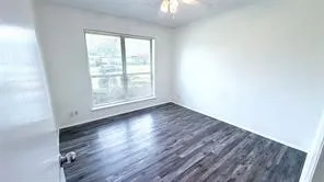 Spacious room featuring wood-finish flooring, a large window, a ceiling fan with light fixture, and white wall finishes