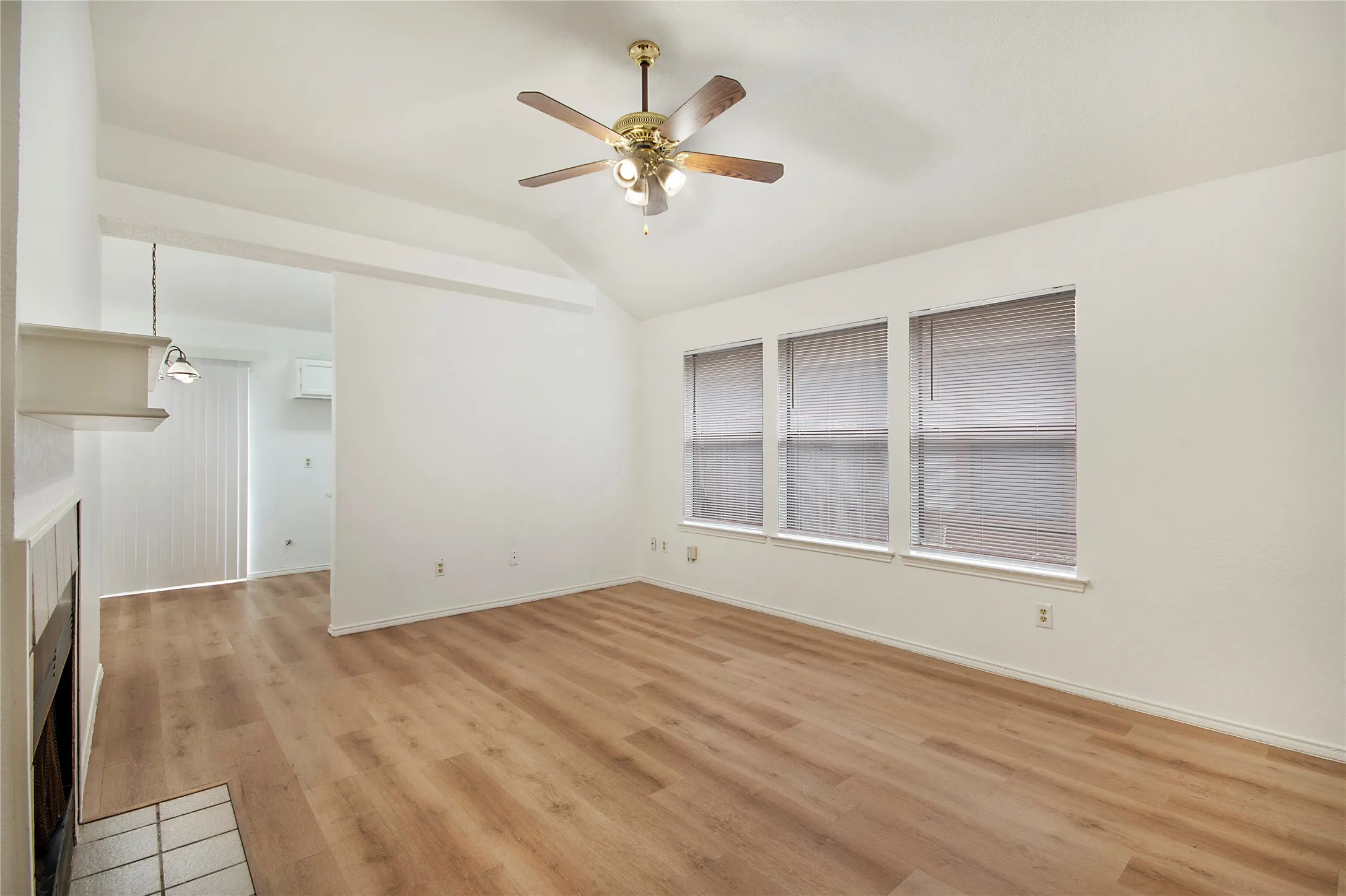 Vaulted ceiling, wood-finish flooring, three large windows, ceiling fan with light fixture, and a built-in fireplace