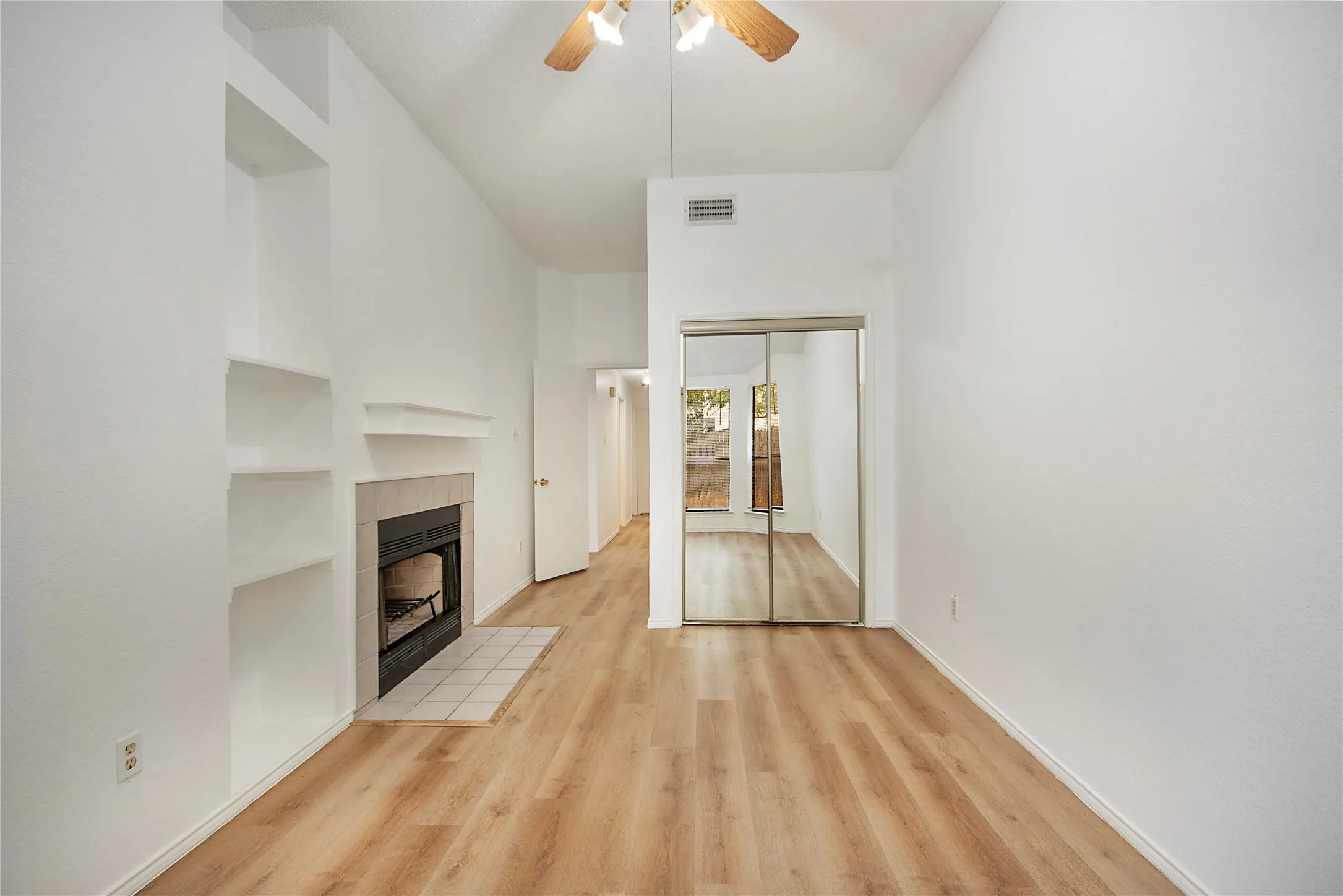 Vaulted ceiling with a ceiling fan, built-in shelving, fireplace with tile surround, wood-finish flooring, and mirrored closet doors
