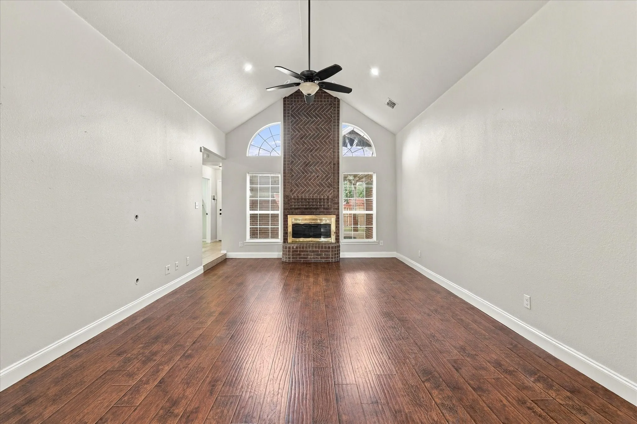 The unique step-down living room is designed to be the gathering space of the home, featuring vaulted ceilings, warm laminate flooring, recessed lighting, ceiling fan and a dramatic floor-to-ceiling brick gas fireplace with raised hearth and wood mantel, beautifully framed by windows.