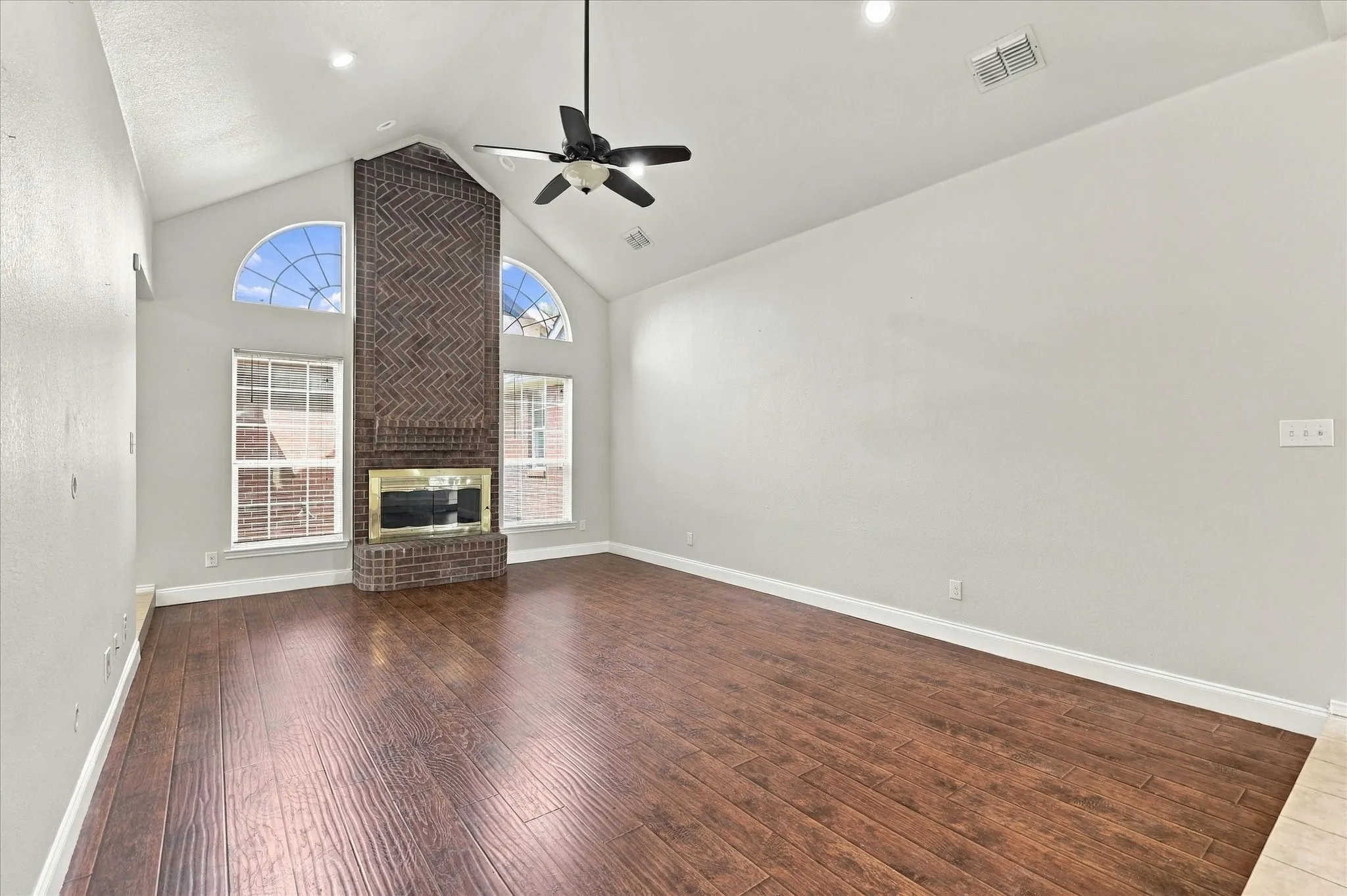 The unique step-down living room is designed to be the gathering space of the home, featuring vaulted ceilings, warm laminate flooring, recessed lighting, ceiling fan and a dramatic floor-to-ceiling brick gas fireplace with raised hearth and wood mantel, beautifully framed by windows.