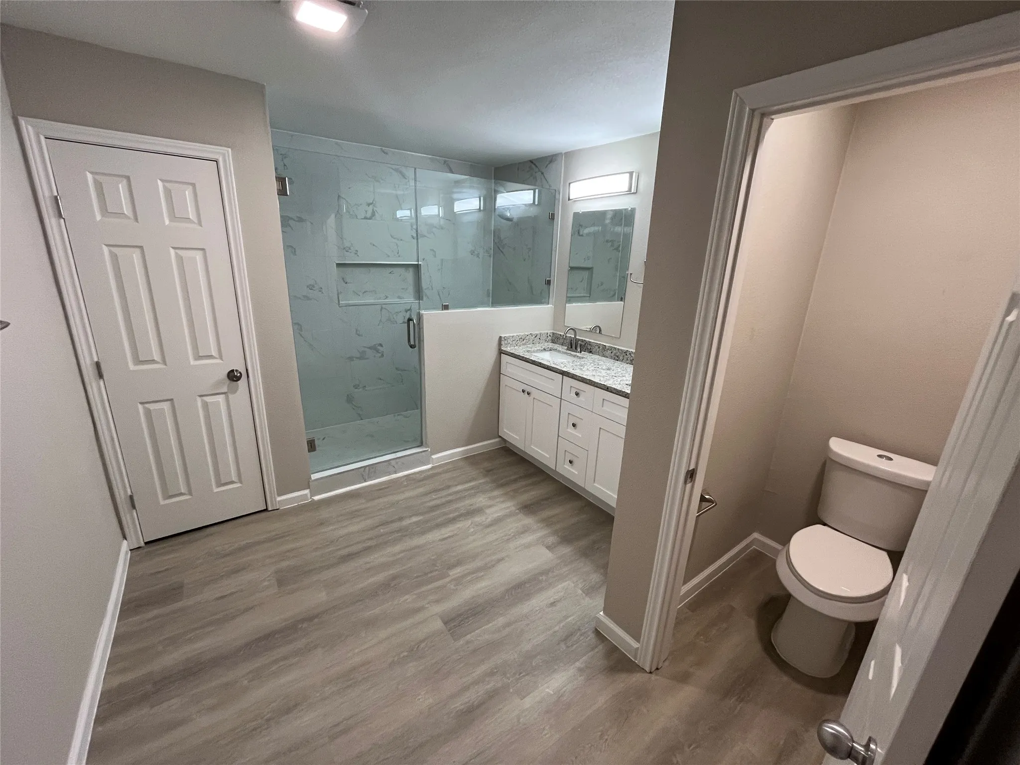 Spacious bathroom featuring wood-finish flooring, a glass-enclosed shower with tile surround, a dual vanity with a light-toned countertop, and recessed lighting