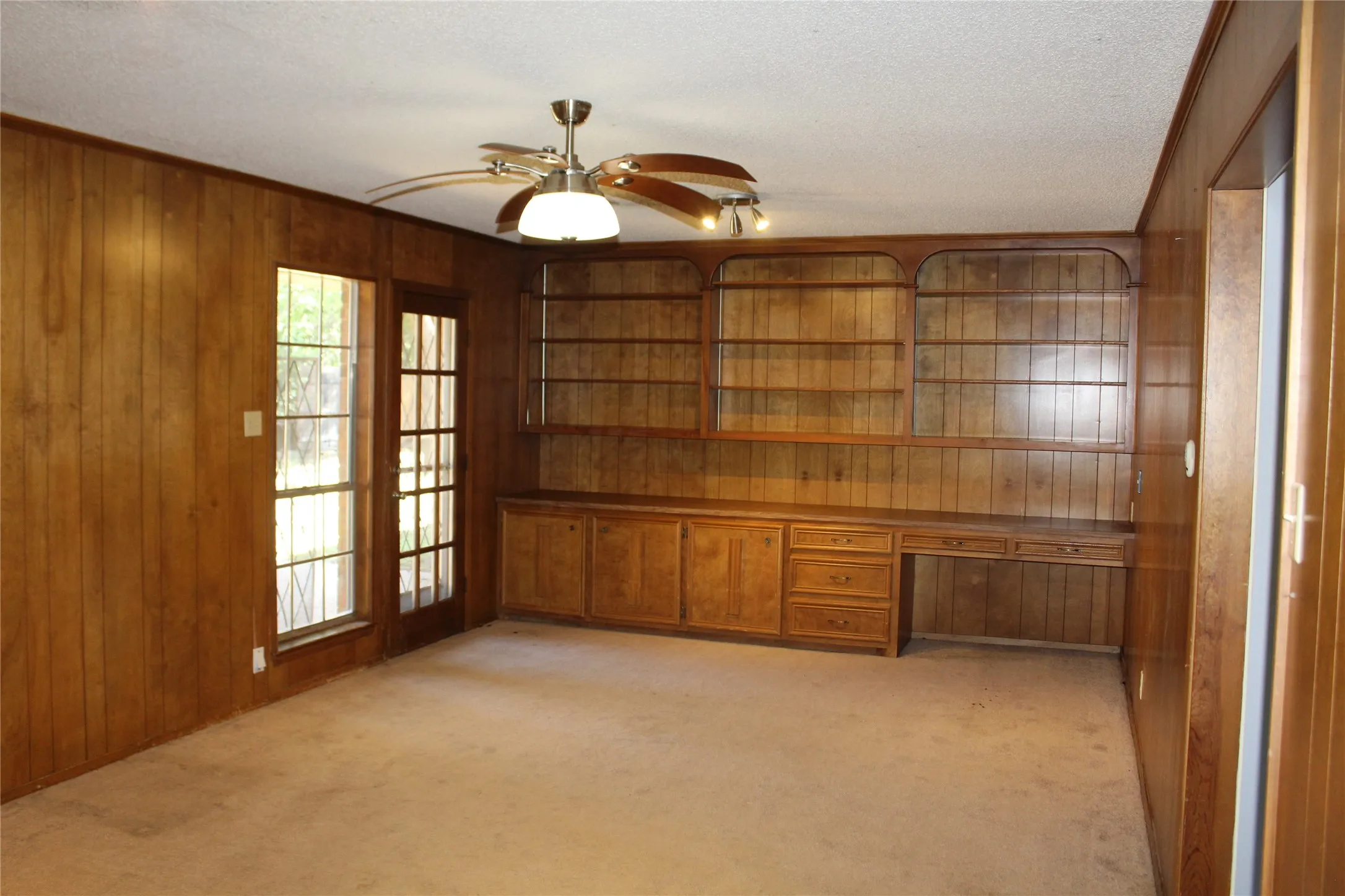Wood paneling throughout, built-in shelving, built-in desk with drawers, glass-paneled door, and a ceiling fan with integrated lighting