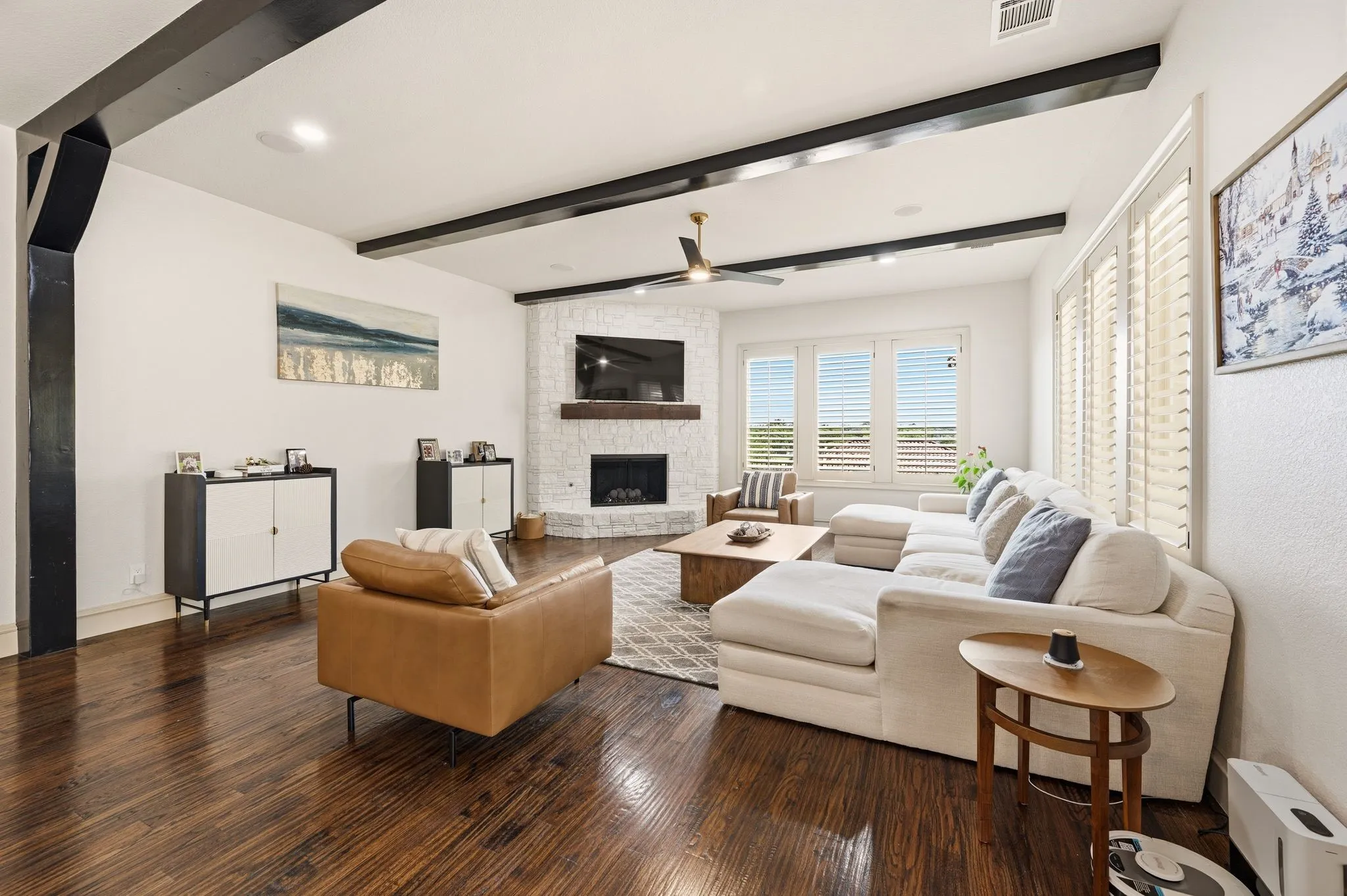 Exposed ceiling beams, wood-finish flooring, corner fireplace with a stone surround, and ceiling fan