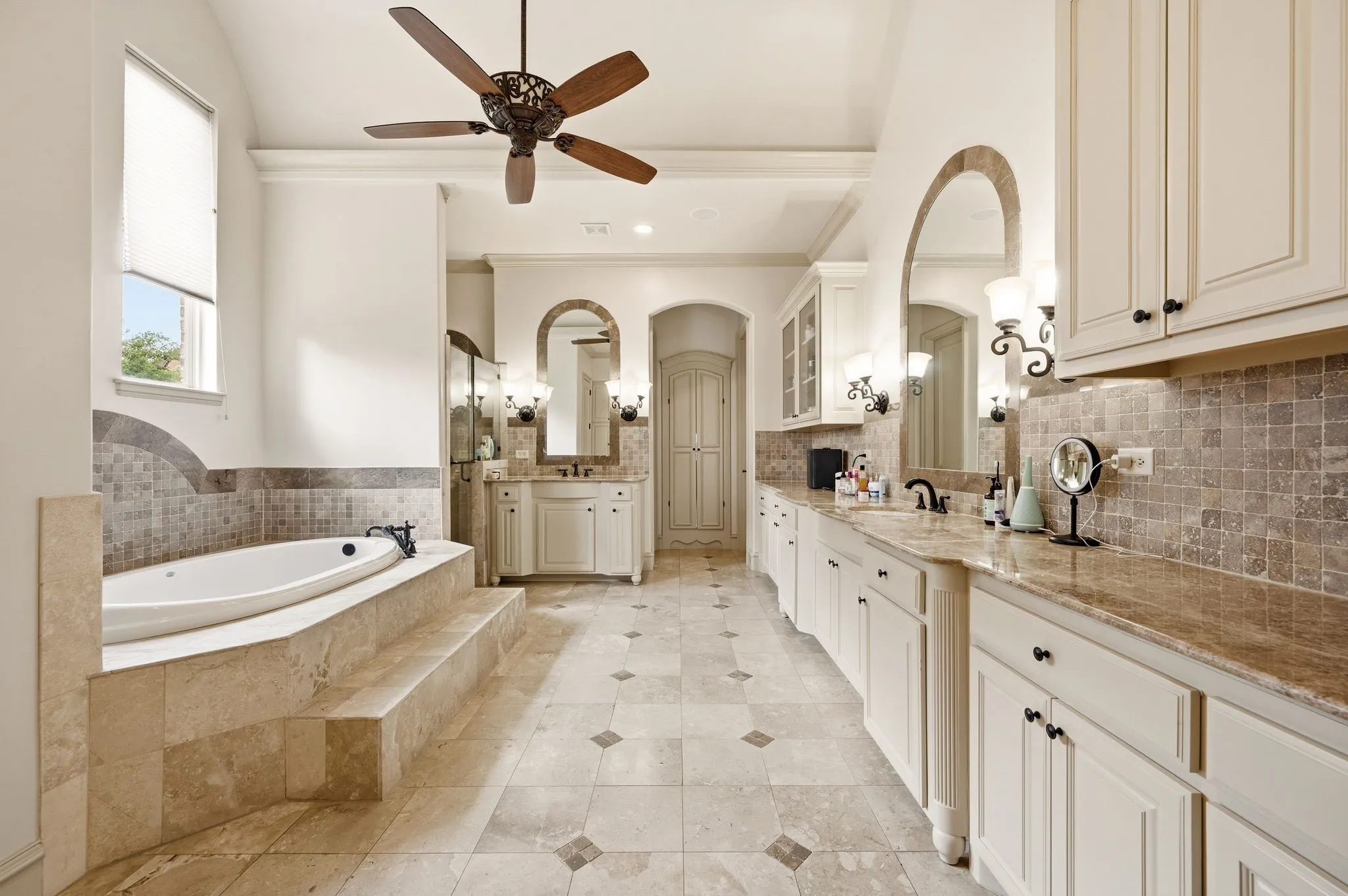 Spacious bathroom featuring a built-in soaking tub, dual vanity areas with stone countertops, tiled flooring, and a decorative ceiling fan