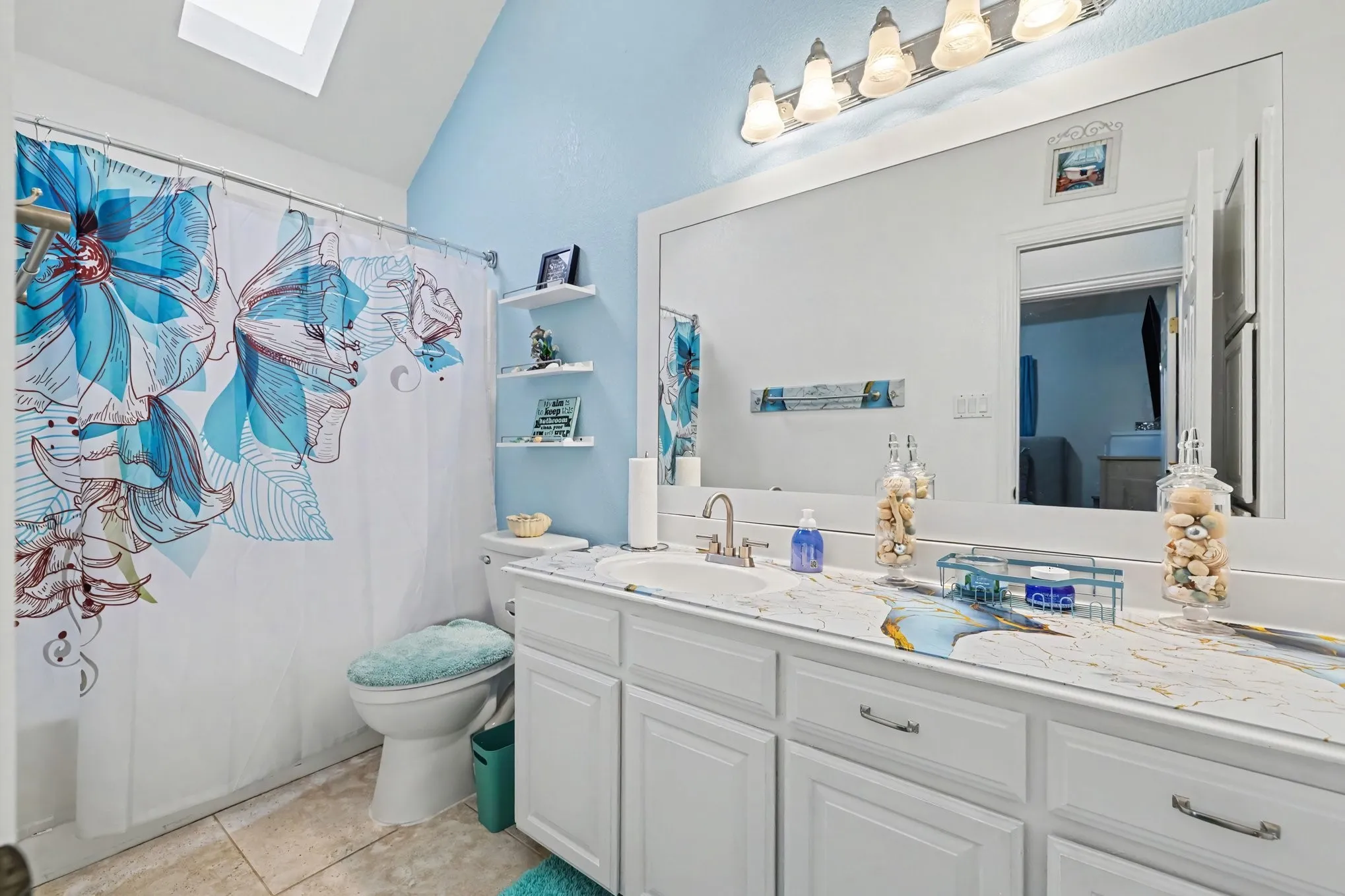 Vaulted ceiling with skylight, dual-sink vanity with white cabinetry, light-toned countertop, and integrated mirror