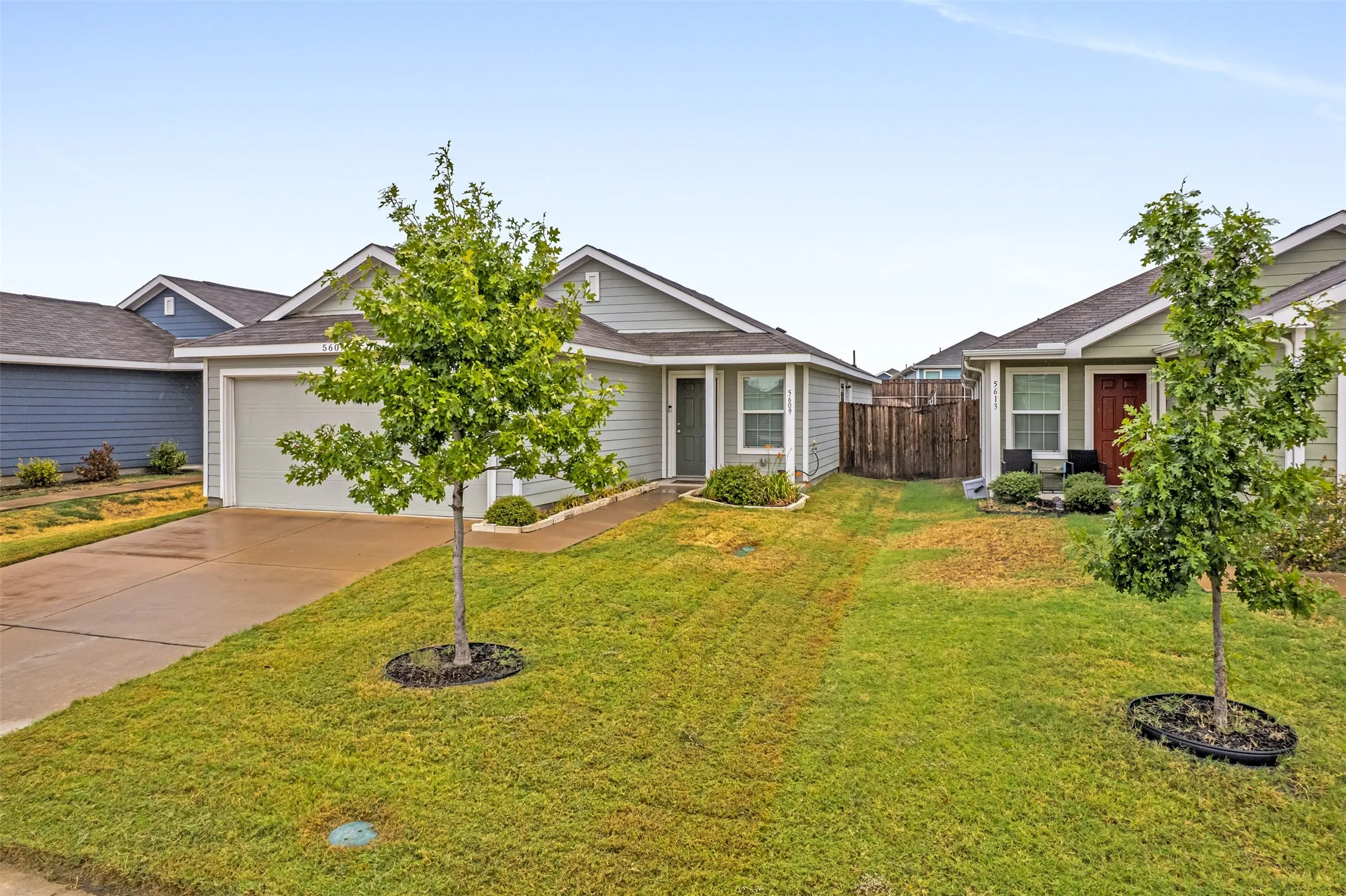 Light-colored siding exterior, attached garage, concrete driveway, front yard with mature trees, and a concrete pathway to the entrance