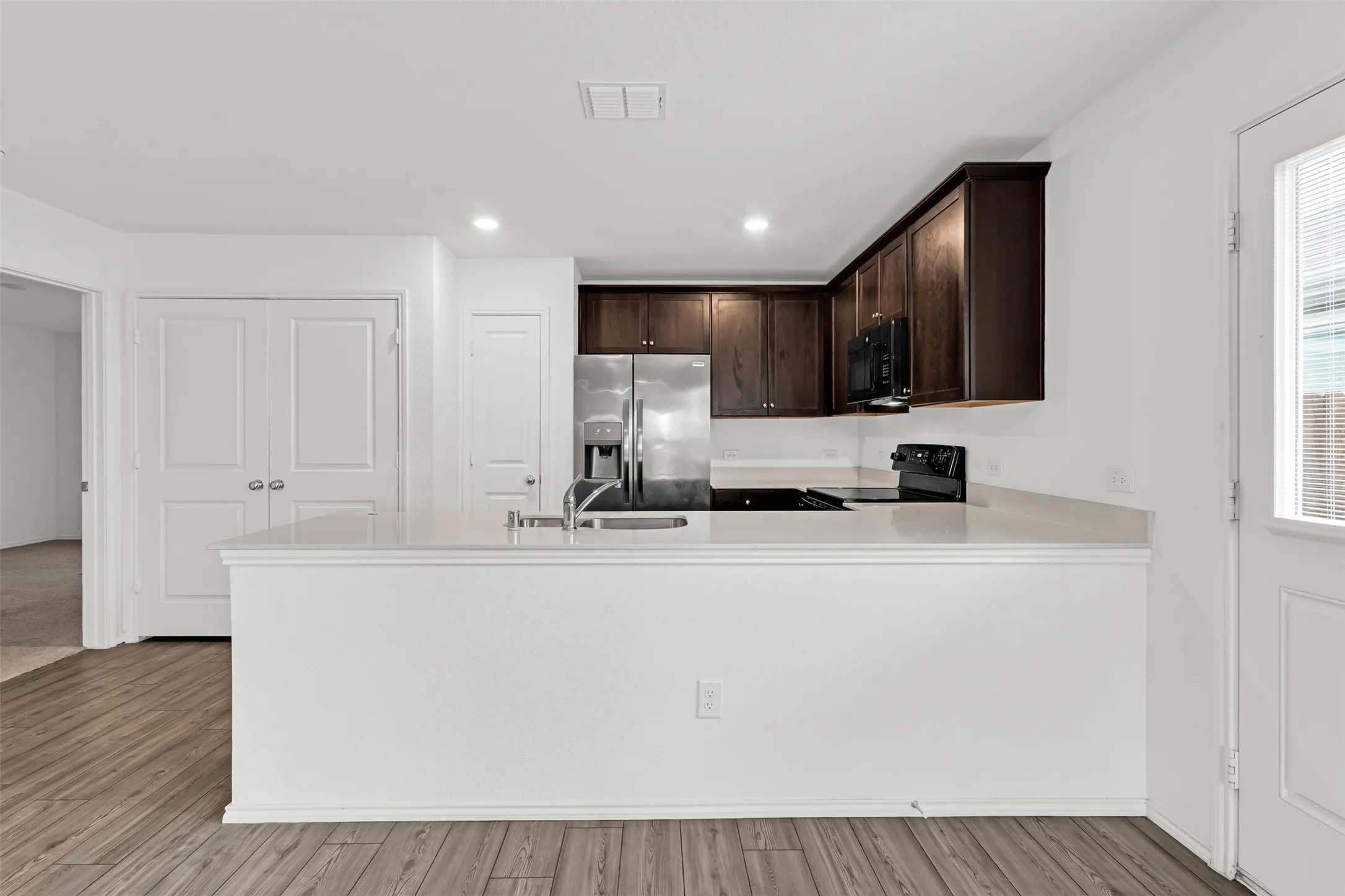 Modern kitchen featuring dark wood cabinetry, stainless steel appliances, quartz countertops, a spacious white island with waterfall edge, recessed lighting, and wide-plank wood-finish flooring throughout the open-concept living area