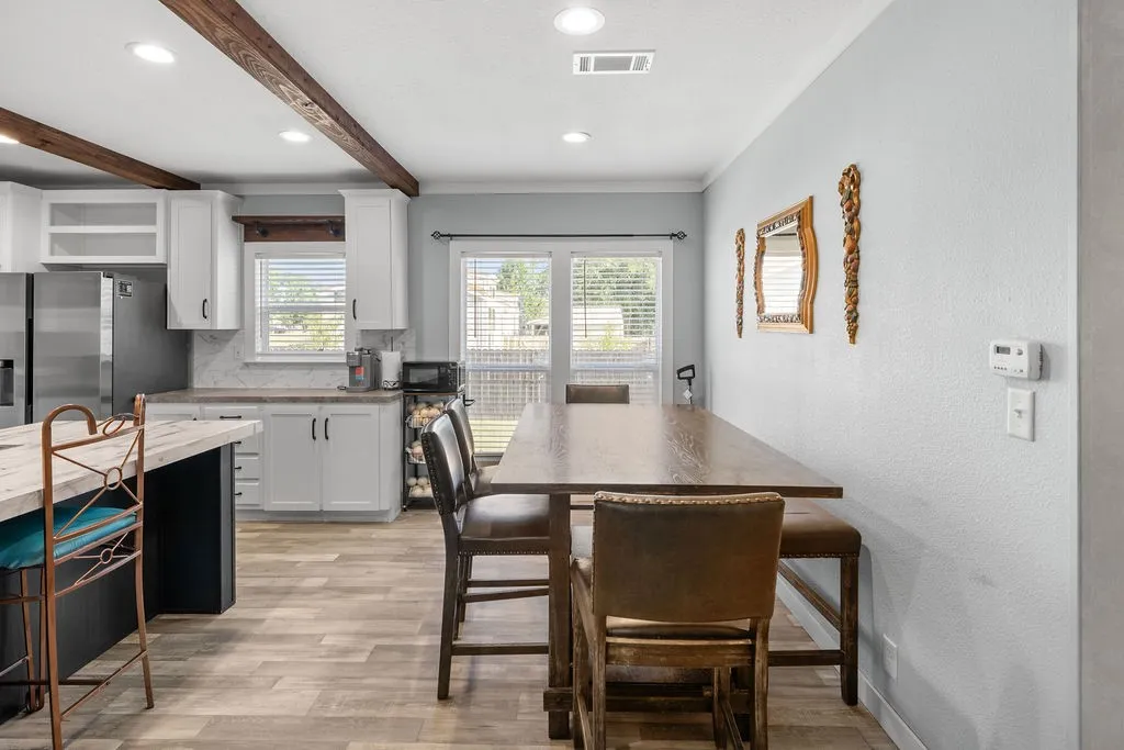 White shaker-style cabinetry with dark hardware, wood-finish flooring, exposed ceiling beams, white tile backsplash, and a sliding glass door