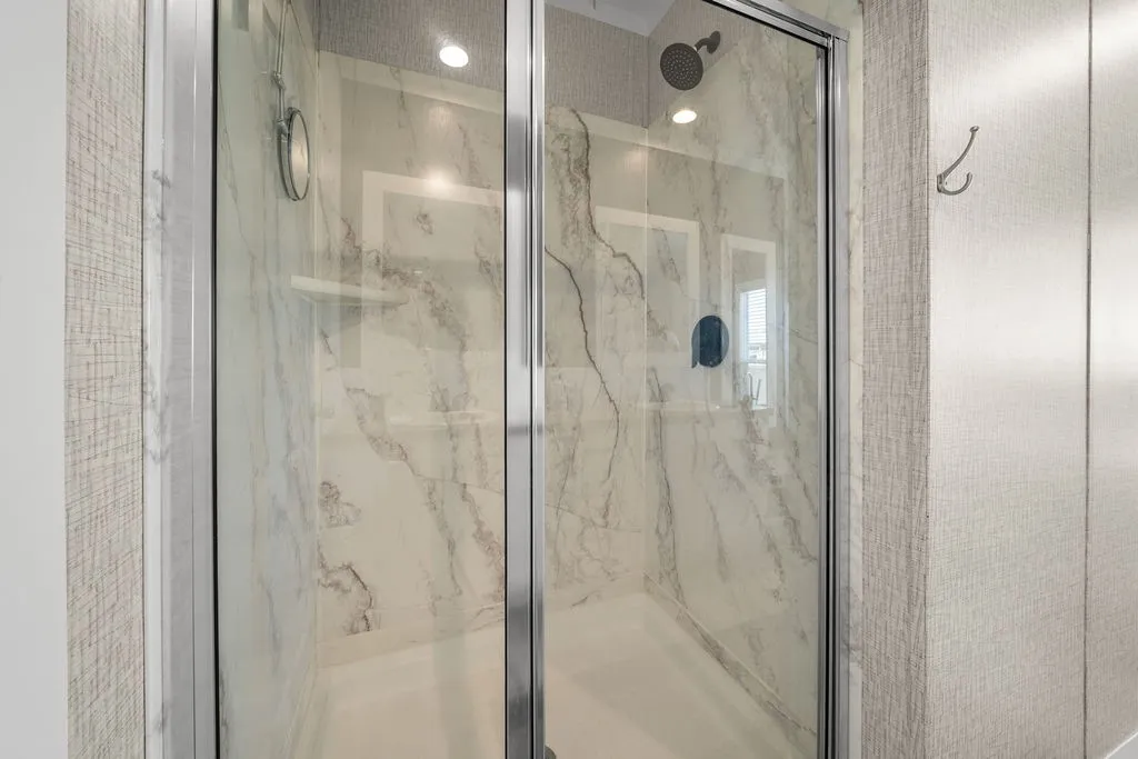 Frameless glass shower enclosure featuring a rain showerhead, built-in shelving, white shower pan, and marble-look wall surround