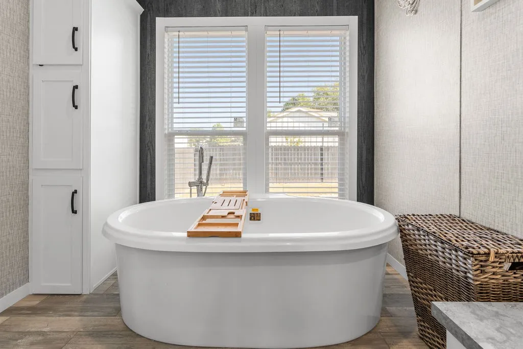 Freestanding soaking tub with a window backdrop, white built-in storage cabinetry, wood-finish flooring, and textured wall coverings