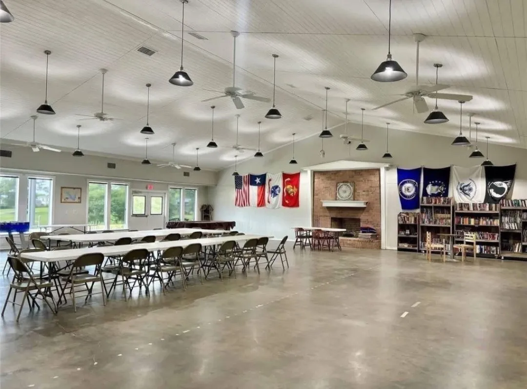 Spacious open hall with a vaulted ceiling, exposed brick fireplace, multiple ceiling fans, pendant lighting, and large windows