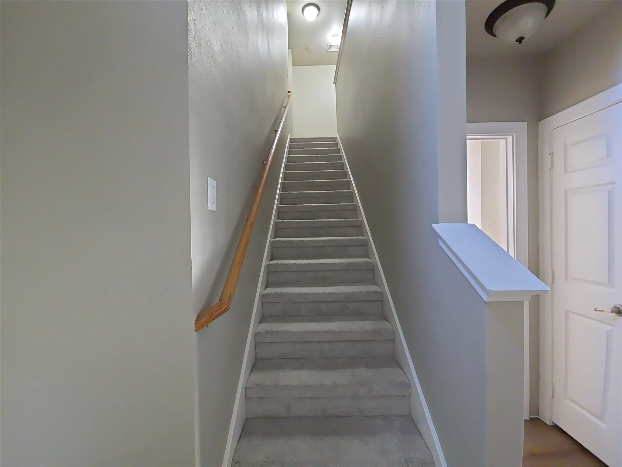Carpeted staircase with a wood handrail, light gray wall finishes, and a flush-mount ceiling light