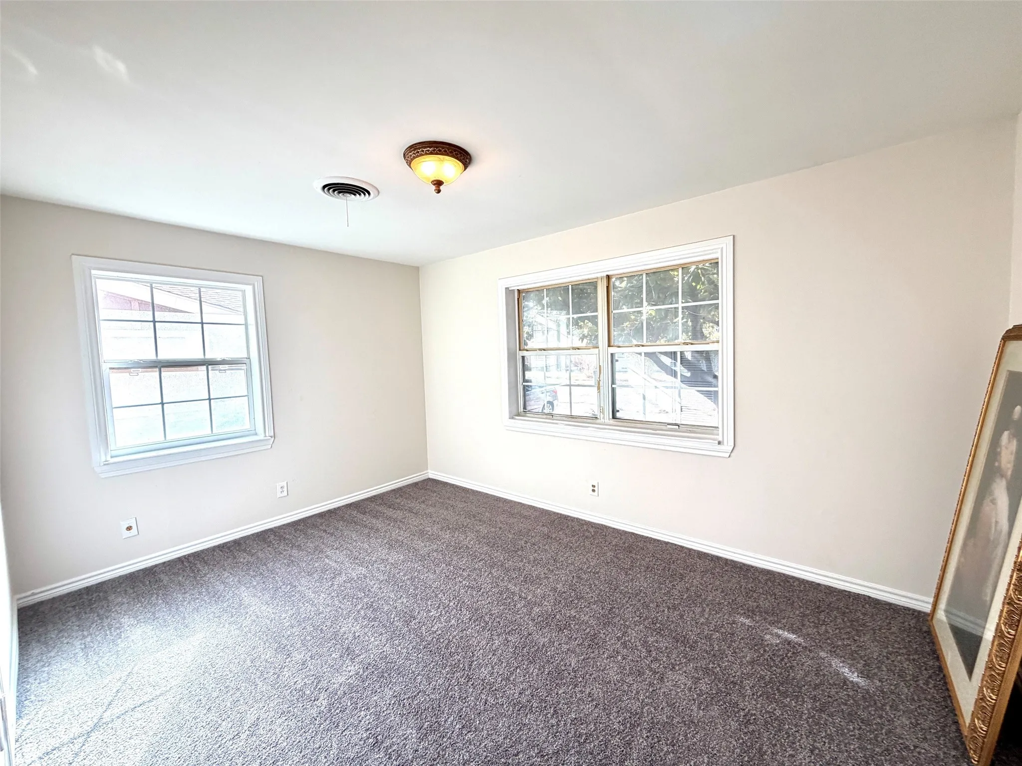 Neutral wall color, two windows with white trim, dark gray carpeting, and a flush-mount ceiling light fixture