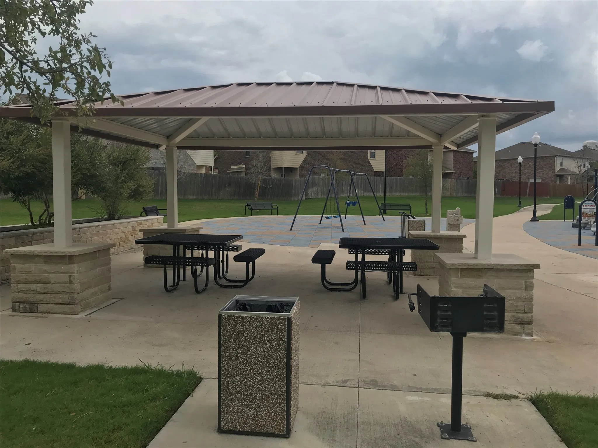 Covered outdoor pavilion with a metal roof and stone-clad support pillars, featuring integrated picnic tables and a built-in grill station