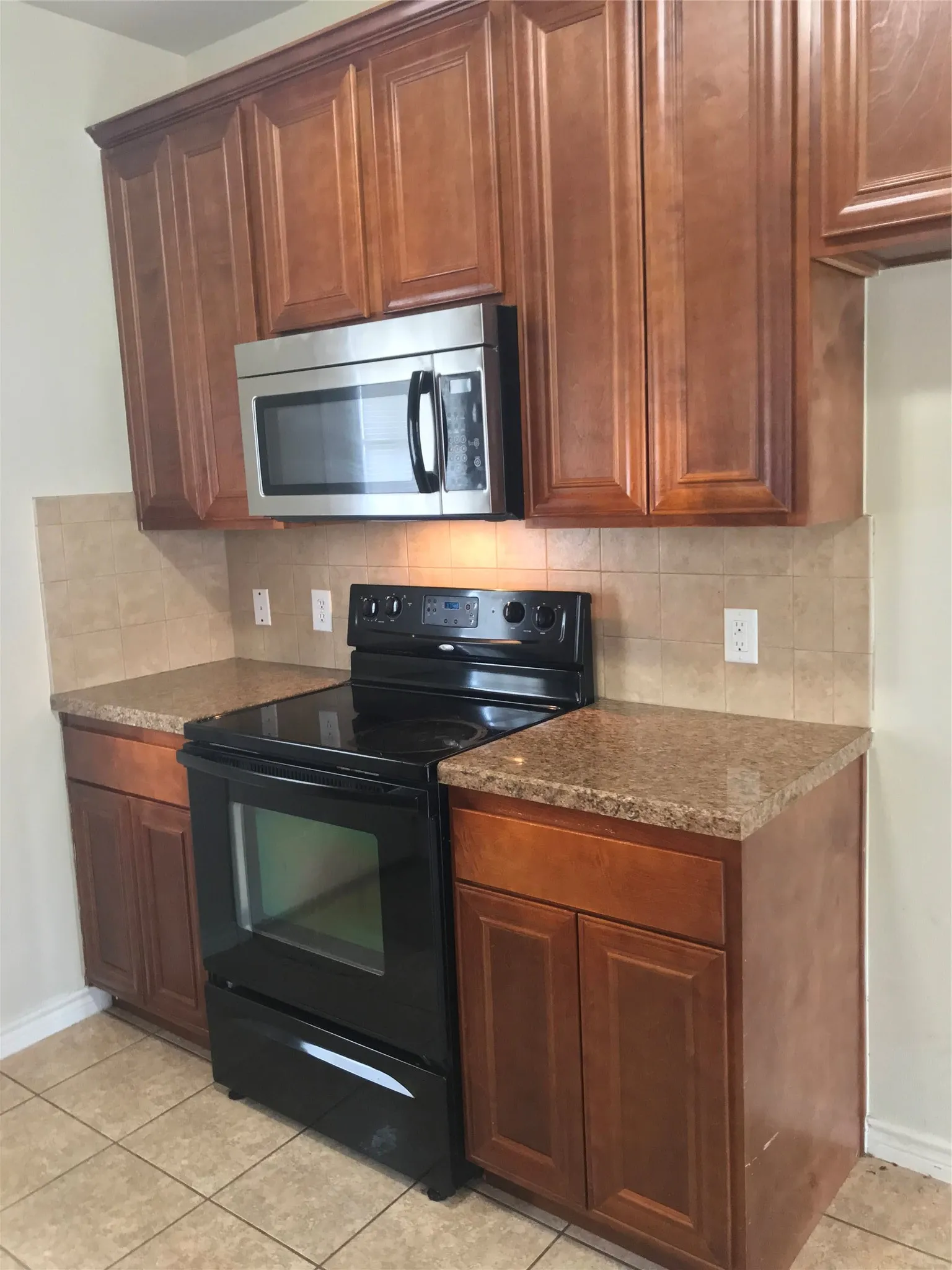 Wood-finish cabinetry complemented by speckled countertops, a tile backsplash, and a stainless steel microwave