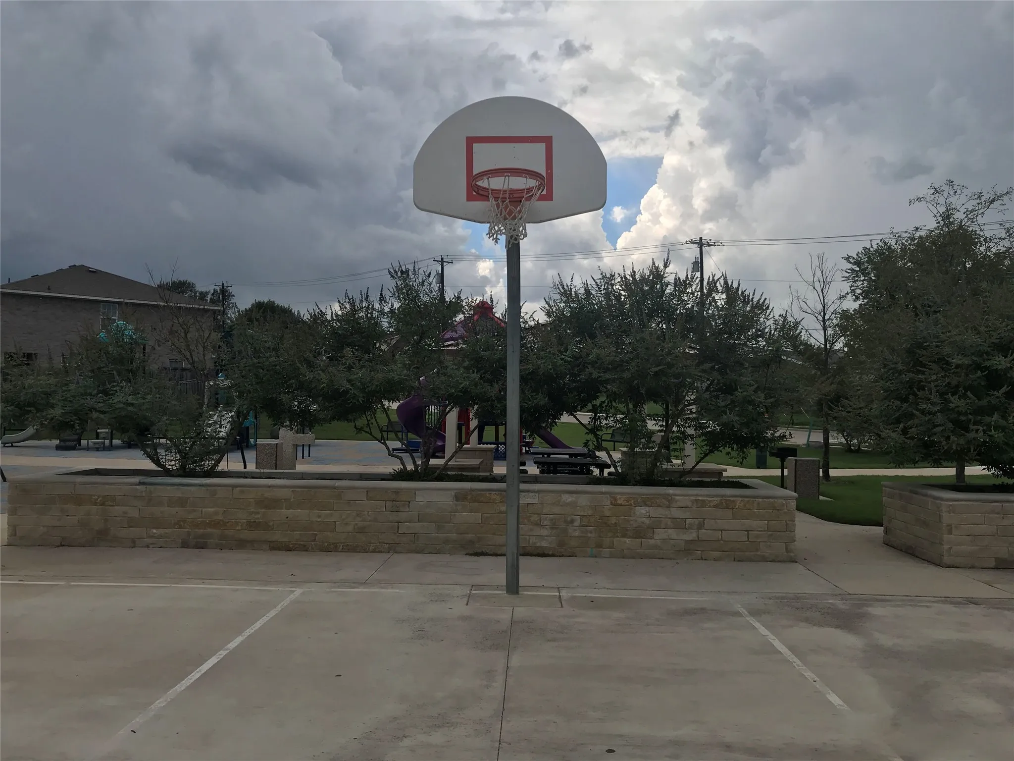 Basketball court featuring a backboard with a red rim, concrete playing surface with painted lines, and stone-clad planters with mature landscaping
