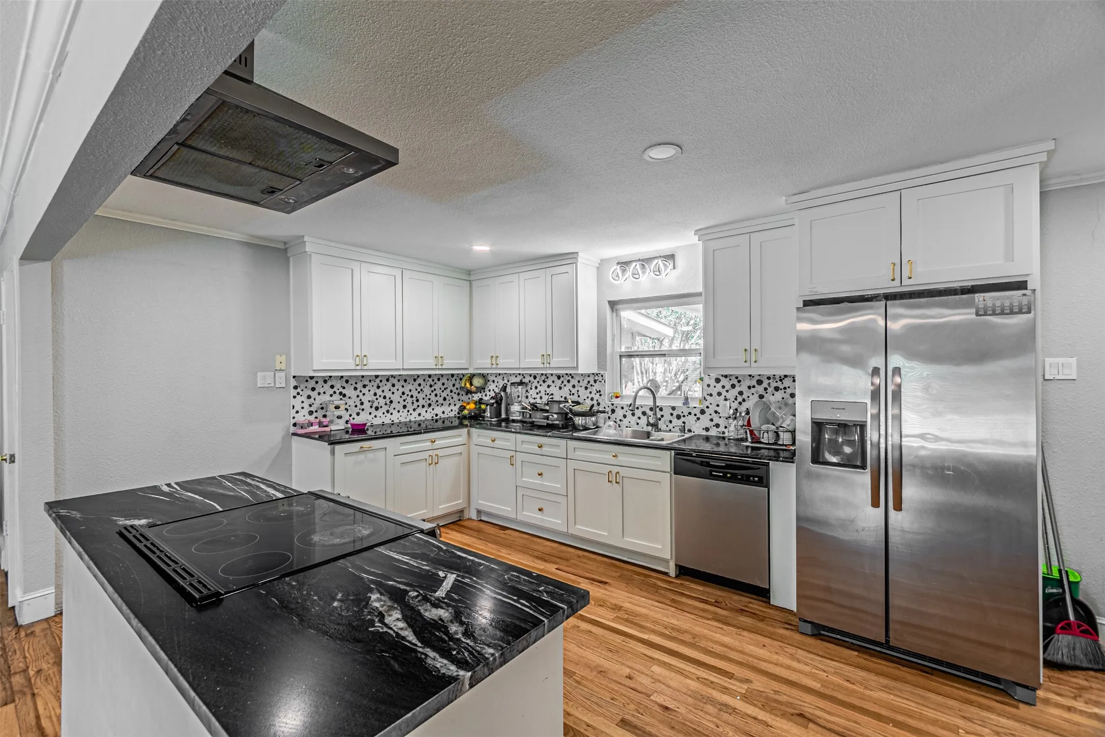 Kitchen featuring black stone countertops, white cabinetry with gold-tone hardware, a built-in cooktop, stainless steel appliances, and wood-finish flooring