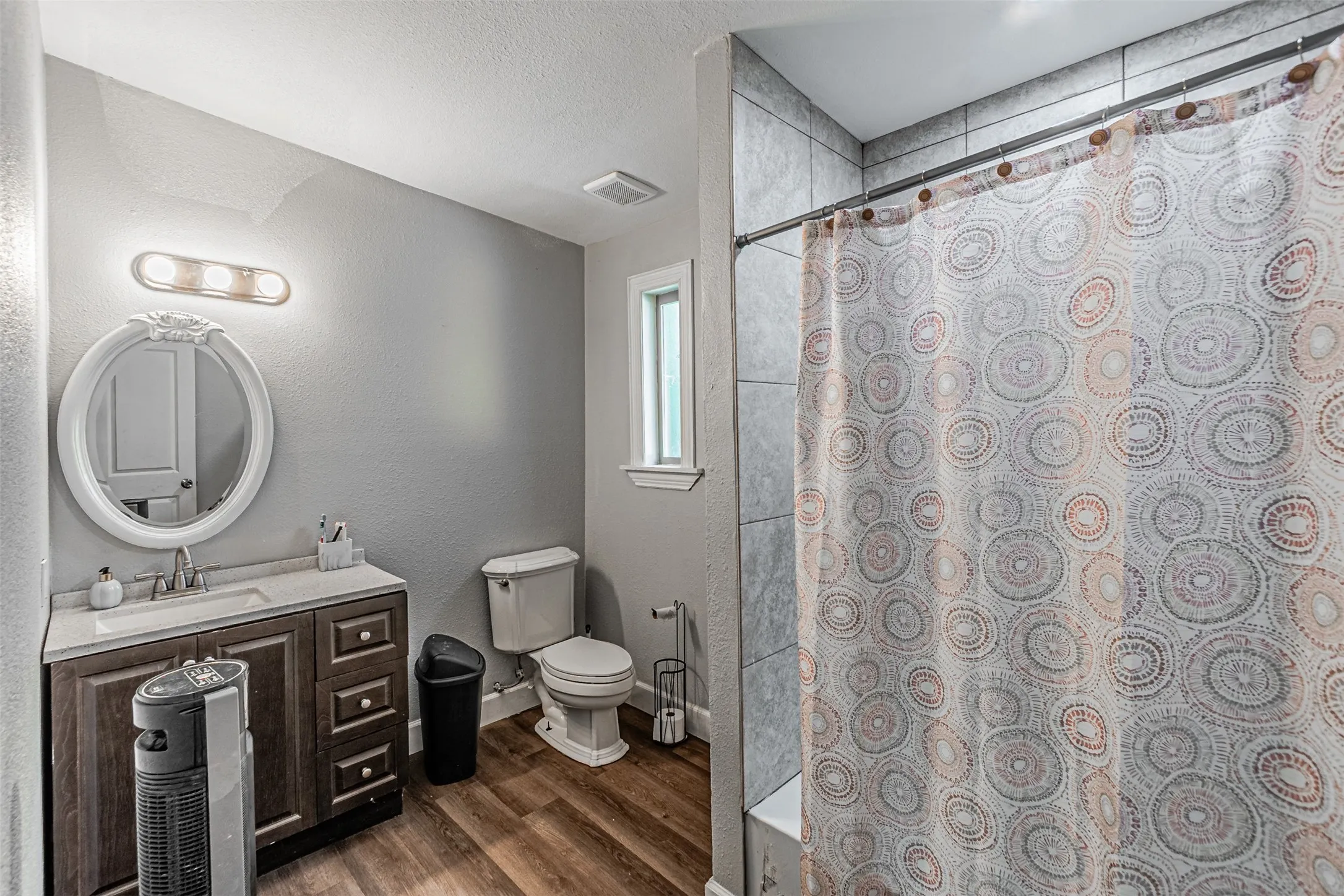 Wood-finish flooring, vanity with a white countertop, oval mirror, wall-mounted light fixture, and tiled shower surround