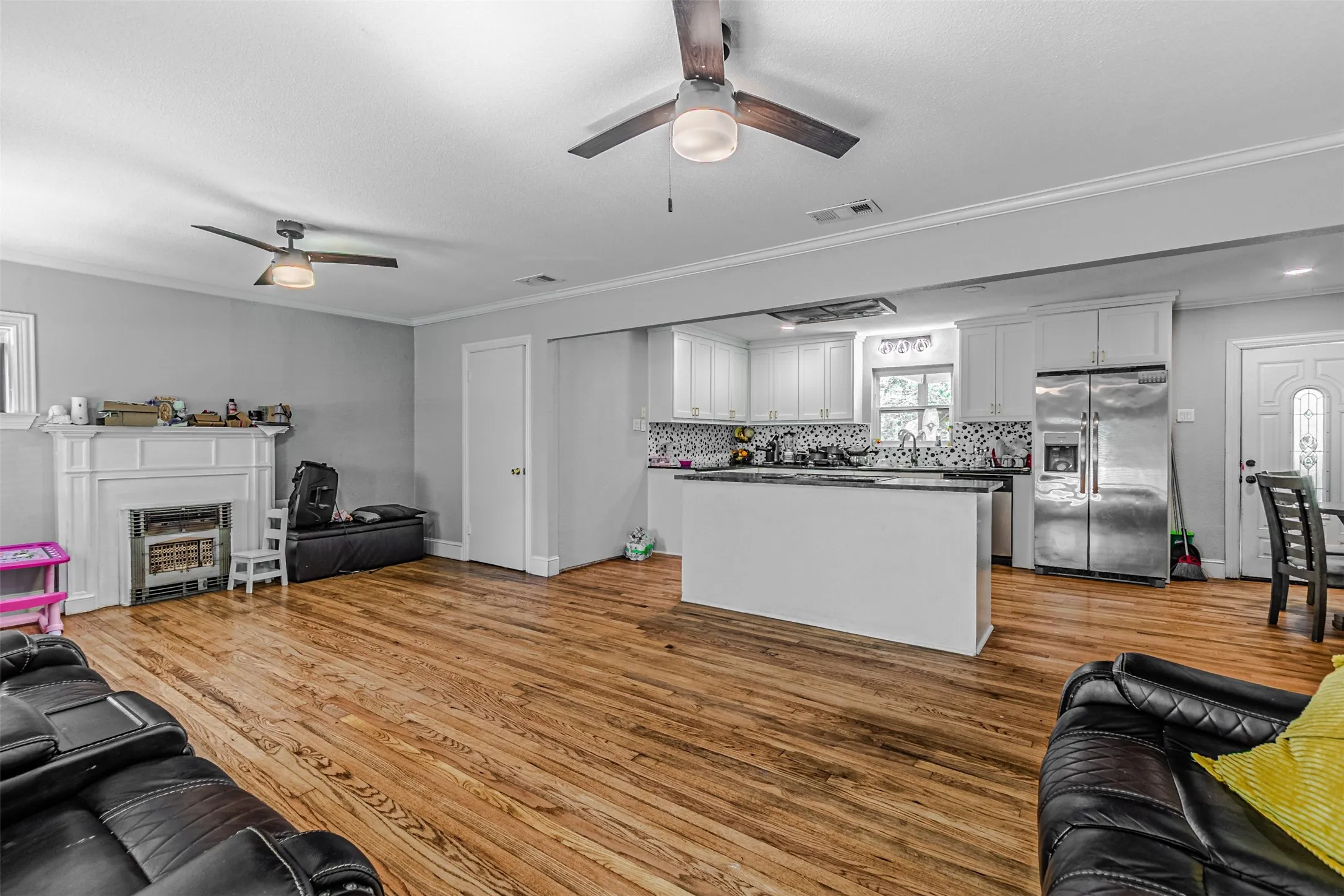 Hardwood flooring, fireplace with mantel, white kitchen cabinetry, stainless steel refrigerator, and ceiling fans