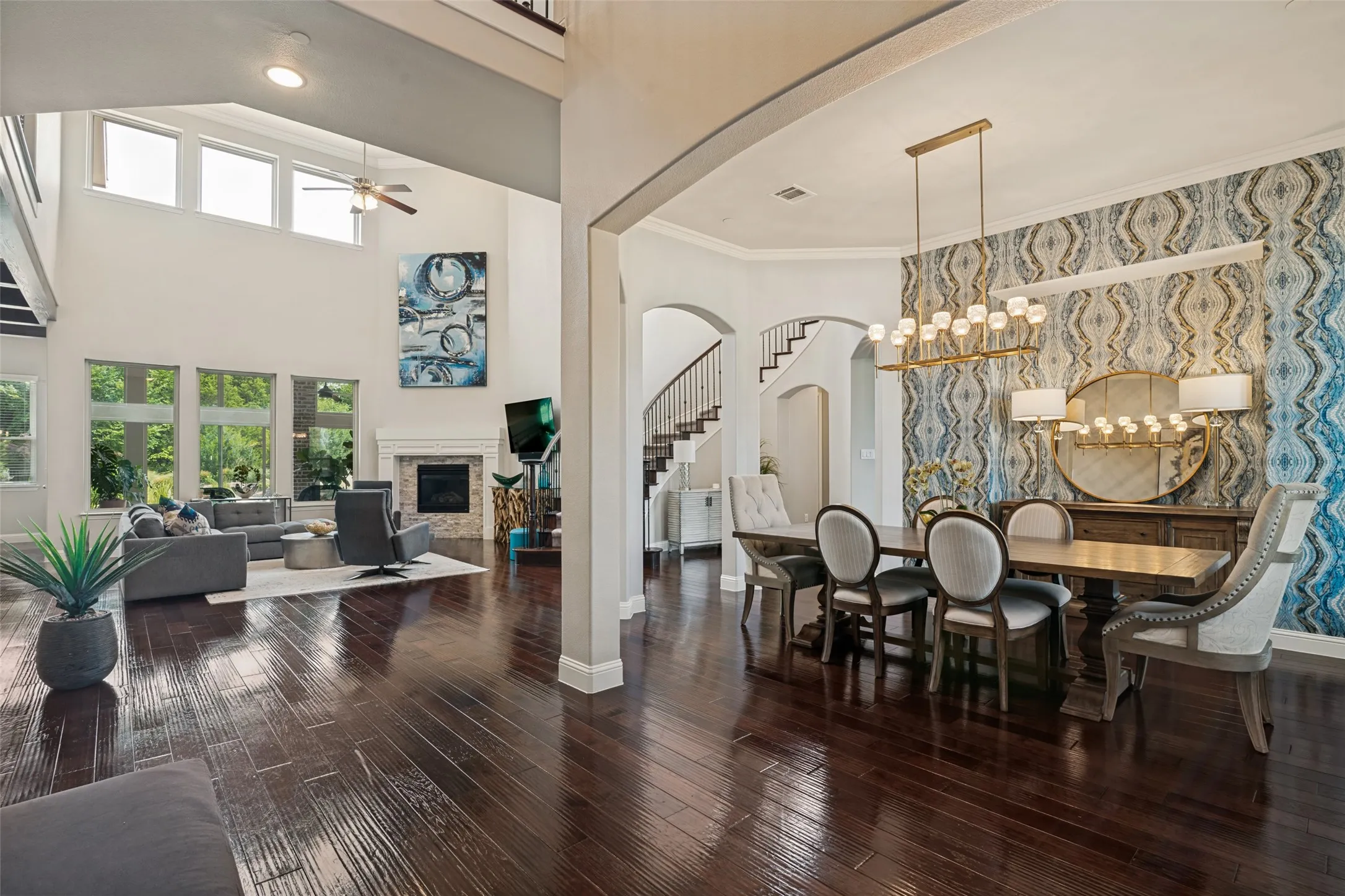 Dark wood-finish flooring throughout, two-story living area with fireplace, expansive windows, arched doorways, and a dining area with a modern chandelier